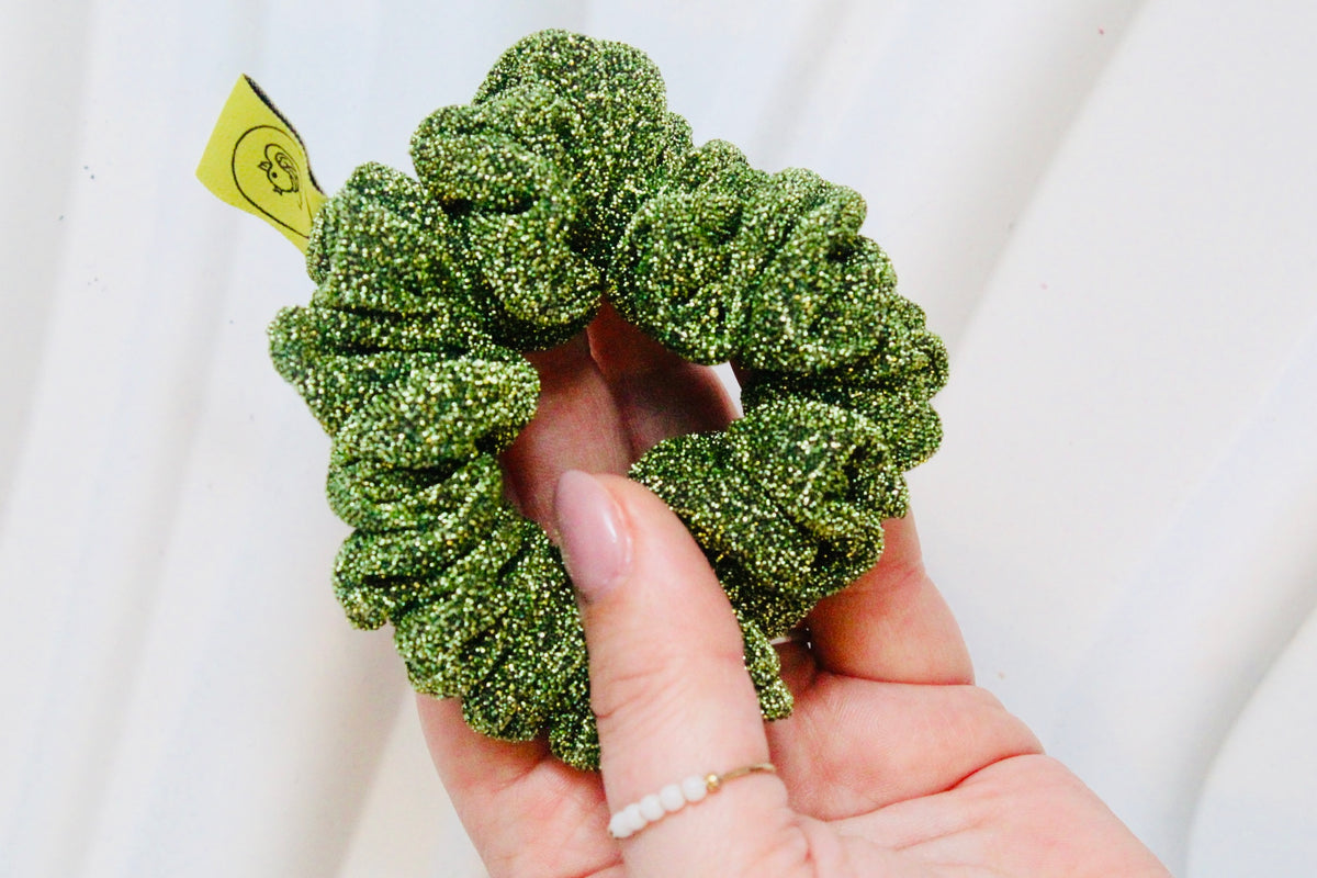 A close-up of a hand holding a green glittery scrunchie with a small yellow tag, set against a soft white draped fabric background.