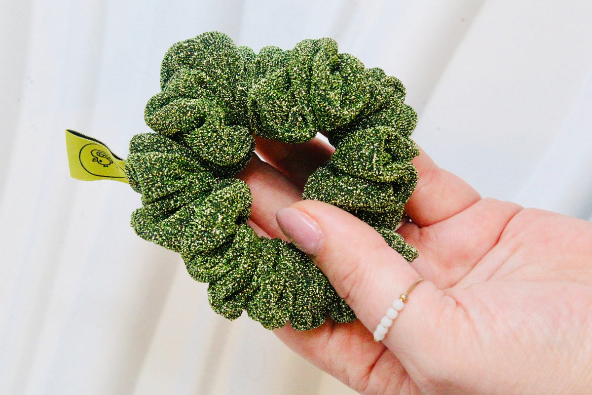 A close-up of a hand holding a green glittery fabric scrunchie with a small yellow tag attached, set against a soft white curtain background.