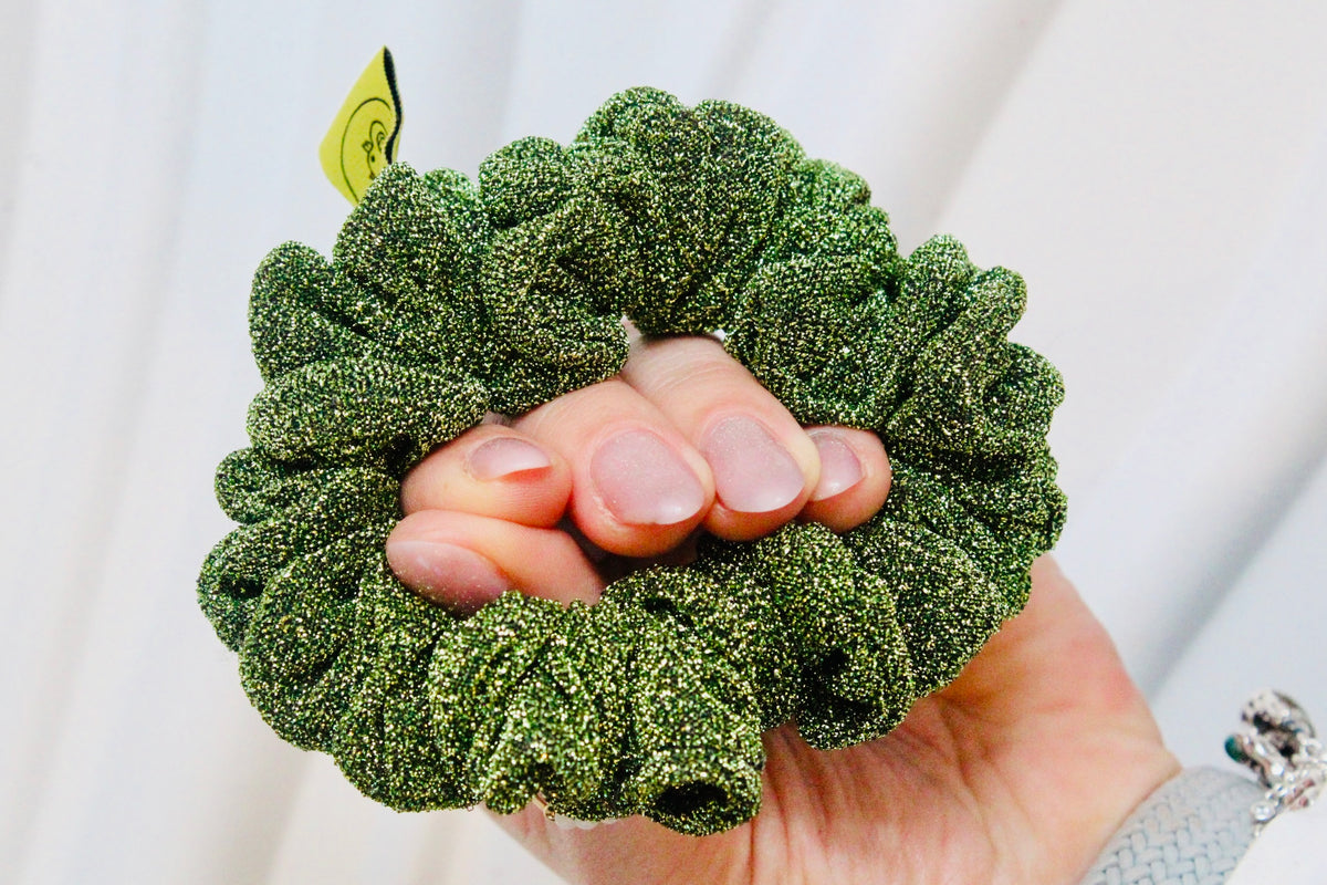A green glittery scrunchie held in a hand against a soft white background.