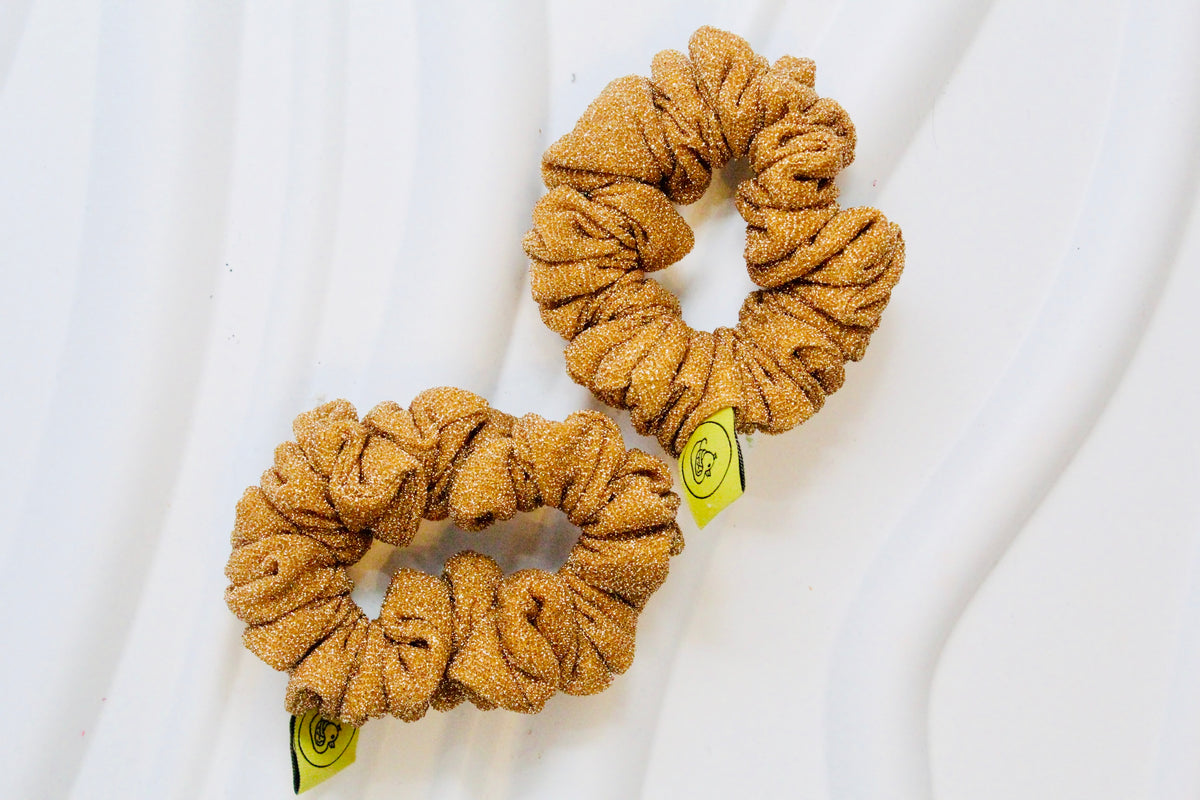 Two textured, ruched scrunchies in a warm mustard-gold color, displayed on a white wavy background with small yellow tags attached.