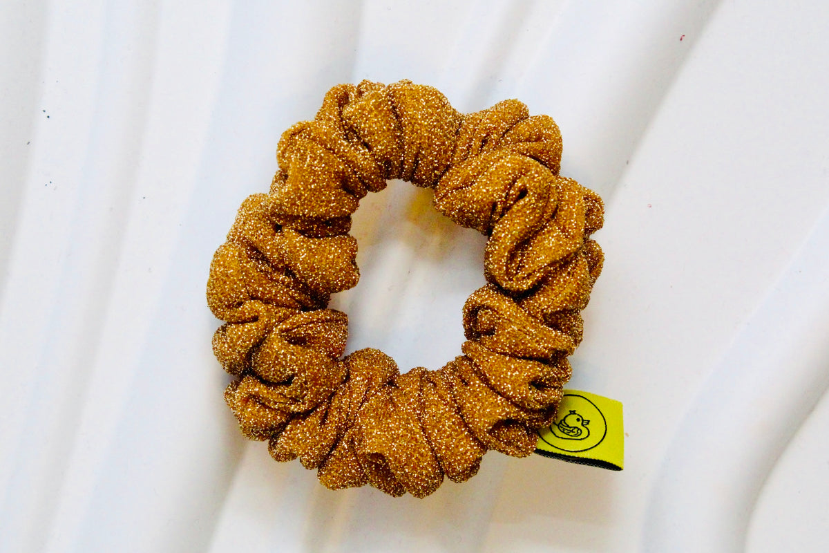 A glittery mustard brown scrunchie with a small yellow tag, displayed on a white textured background.