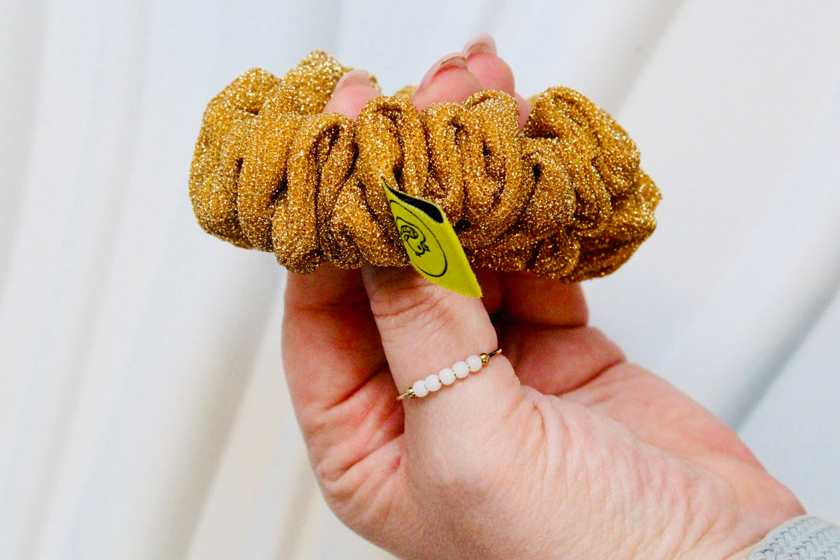 A hand holding a gold glitter scrunchie with a small yellow tag, set against a soft white draped fabric background.