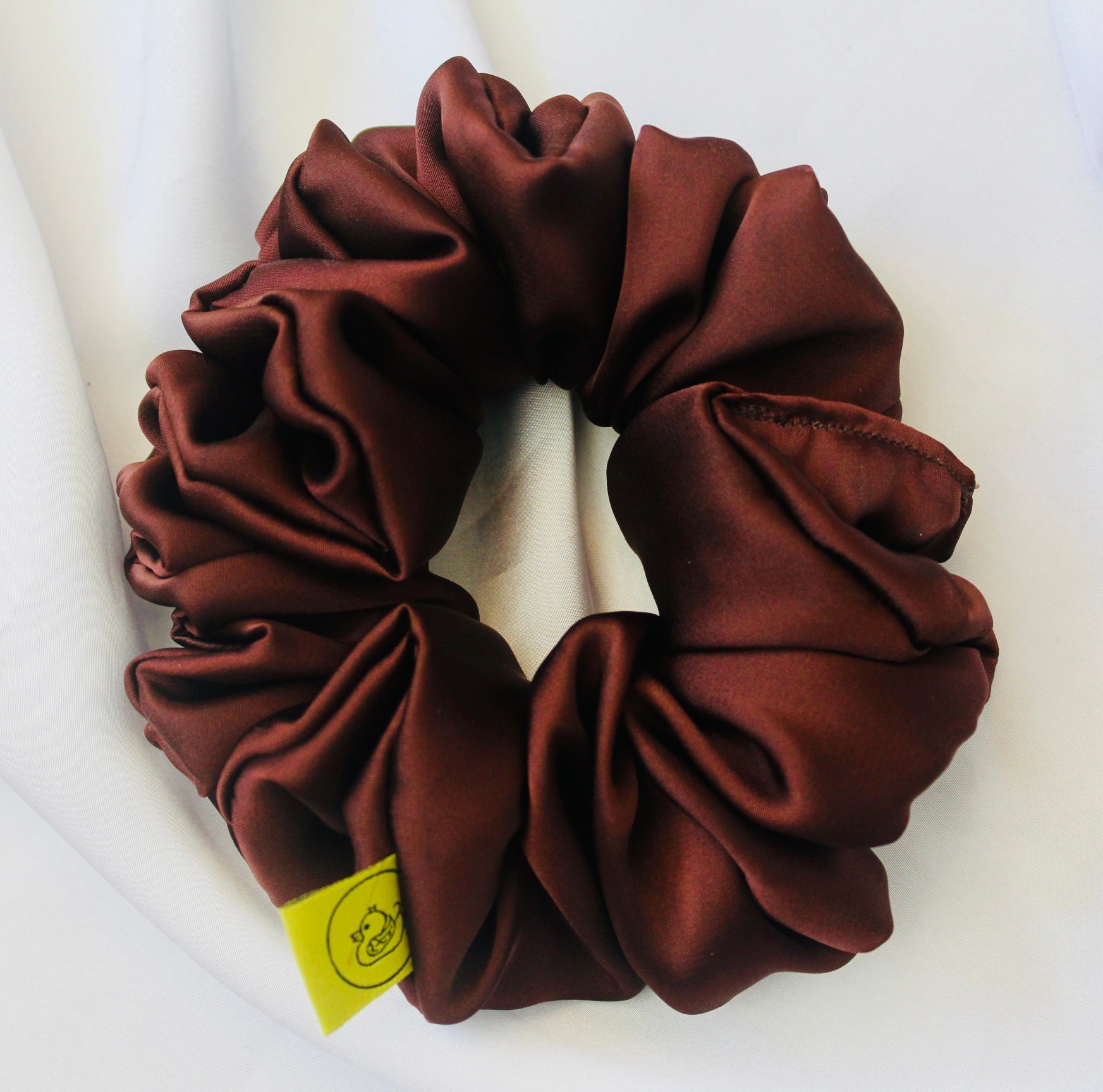 A satin scrunchie in a rich chocolate brown shade with a smooth and shiny texture and a ruched design.  This scrunchie is made from glossy satin giving it a soft, luxurious touch.  The deep chocolate brown colour is both bold and neutral, making it easy to style with a wide range of outfits.  The fabric is softly ruched over elastic, creating volume while remaining comfortable to wear.