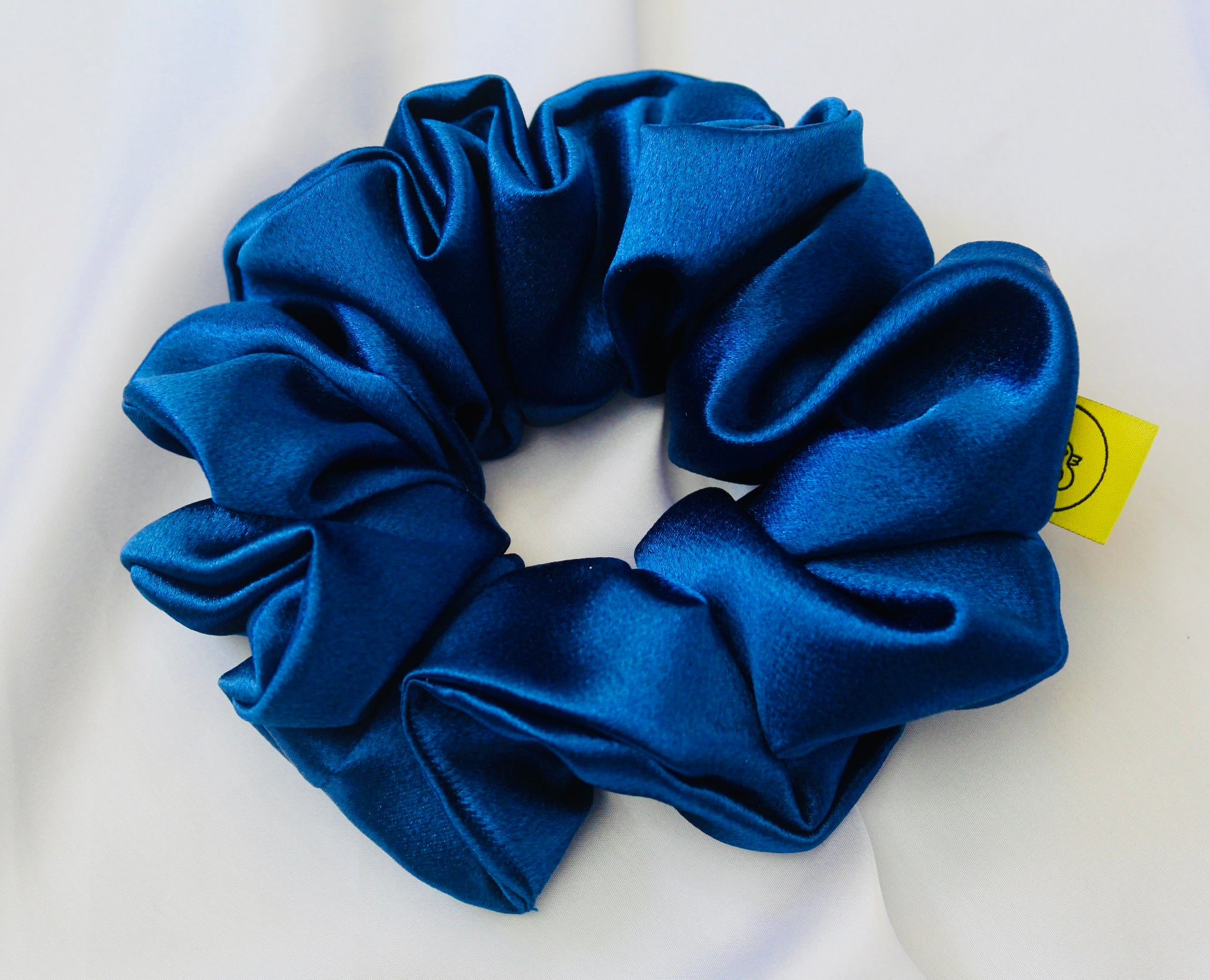 A satin scrunchie in a striking royal blue shade with a silky glossy texture and a ruched design.  Crafted from satin this scrunchie feels smooth and cool to the touch.  The bold royal blue colour stands out beautifully, making it a statement piece for both casual and evening looks.  The gathered satin fabric creates a full textured effect over the elastic ensuring a secure hold.