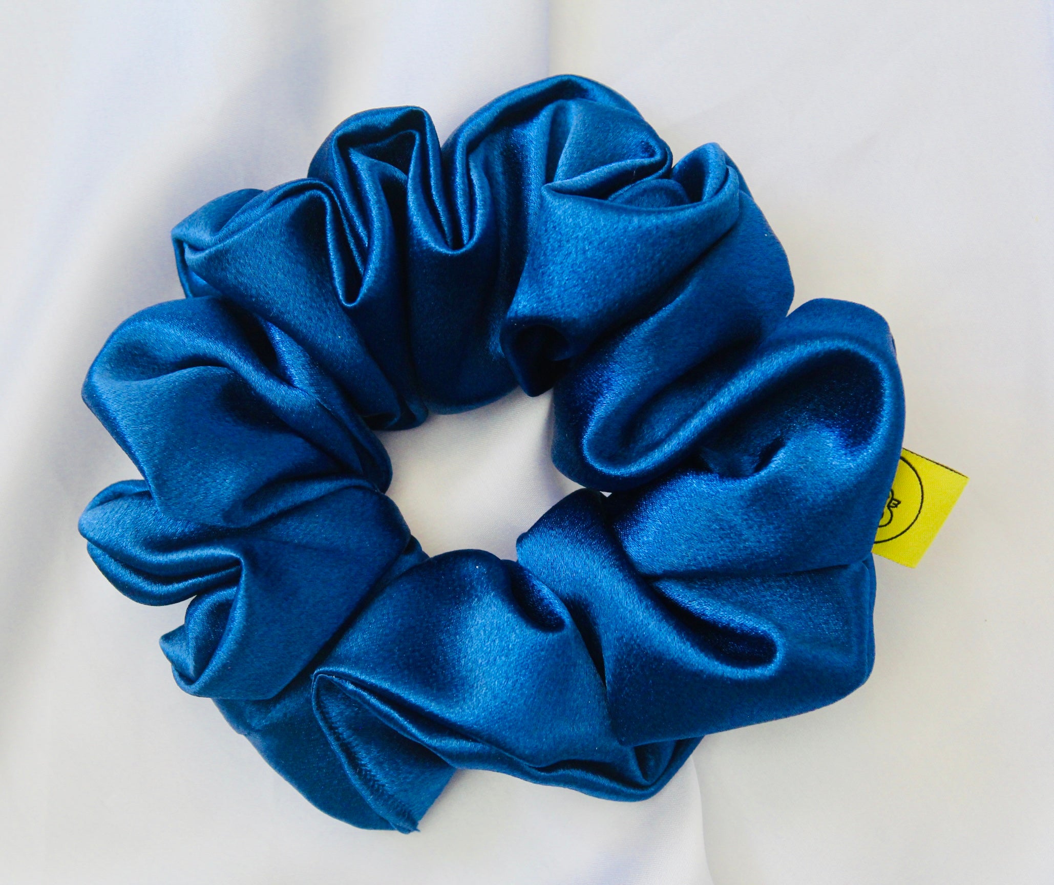 A satin scrunchie in a striking royal blue shade with a silky glossy texture and a ruched design.  Crafted from satin this scrunchie feels smooth and cool to the touch.  The bold royal blue colour stands out beautifully, making it a statement piece for both casual and evening looks.  The gathered satin fabric creates a full textured effect over the elastic ensuring a secure hold.