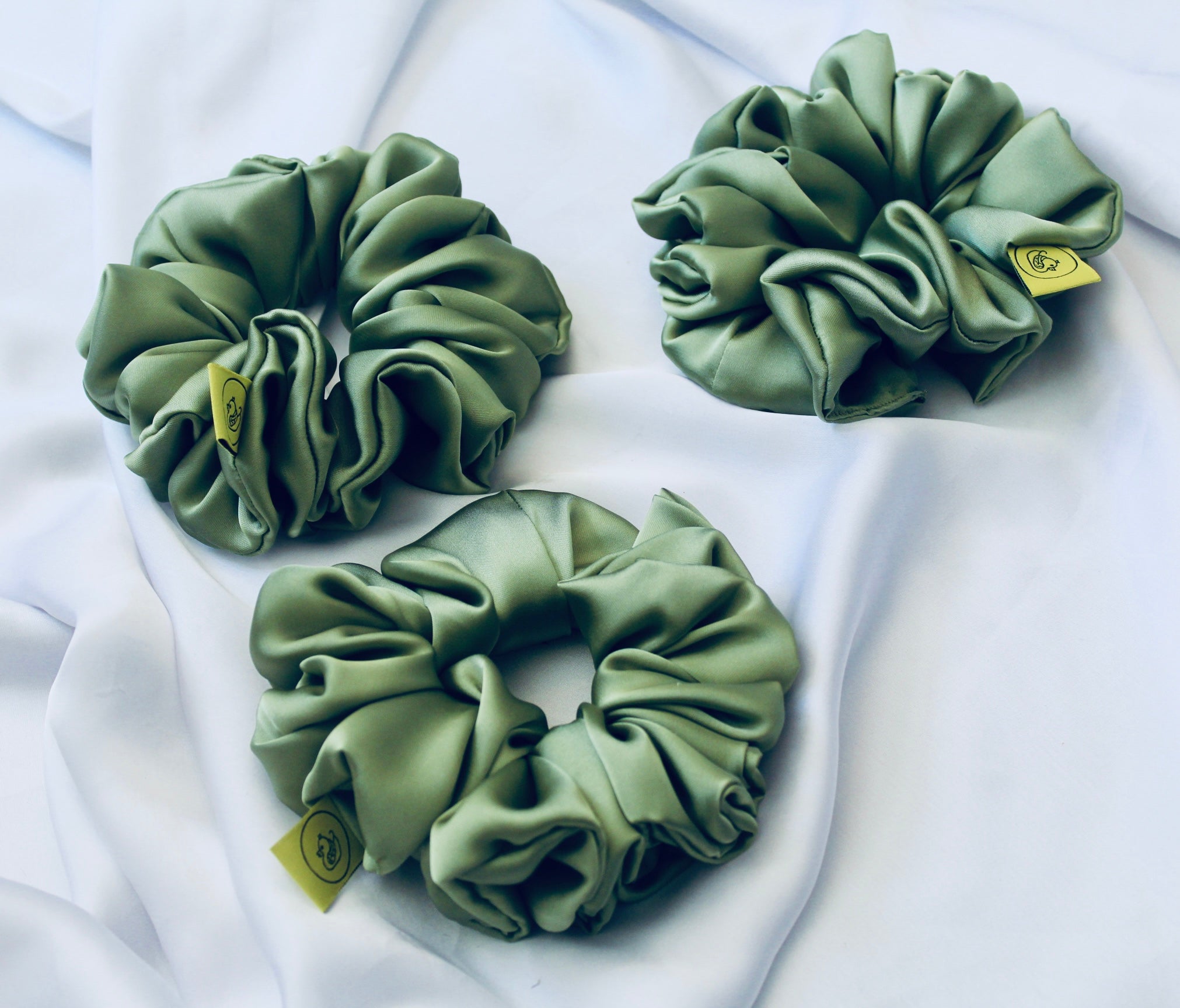 A satin scrunchie in a light green shade with a silky, glossy finish and a ruched design.  Crafted from smooth satin, this scrunchie feels soft and sleek against the hair.  The green tone has a timeless, earthy quality, perfect for both everyday wear and festive styling.  Its ruched fabric is stretched over an elastic band, providing a secure yet comfortable fit.