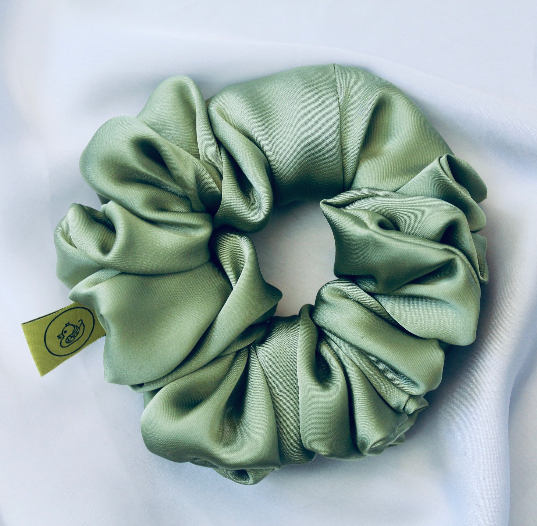 A satin scrunchie in a light green shade with a silky, glossy finish and a ruched design.  Crafted from smooth satin, this scrunchie feels soft and sleek against the hair.  The green tone has a timeless, earthy quality, perfect for both everyday wear and festive styling.  Its ruched fabric is stretched over an elastic band, providing a secure yet comfortable fit.
