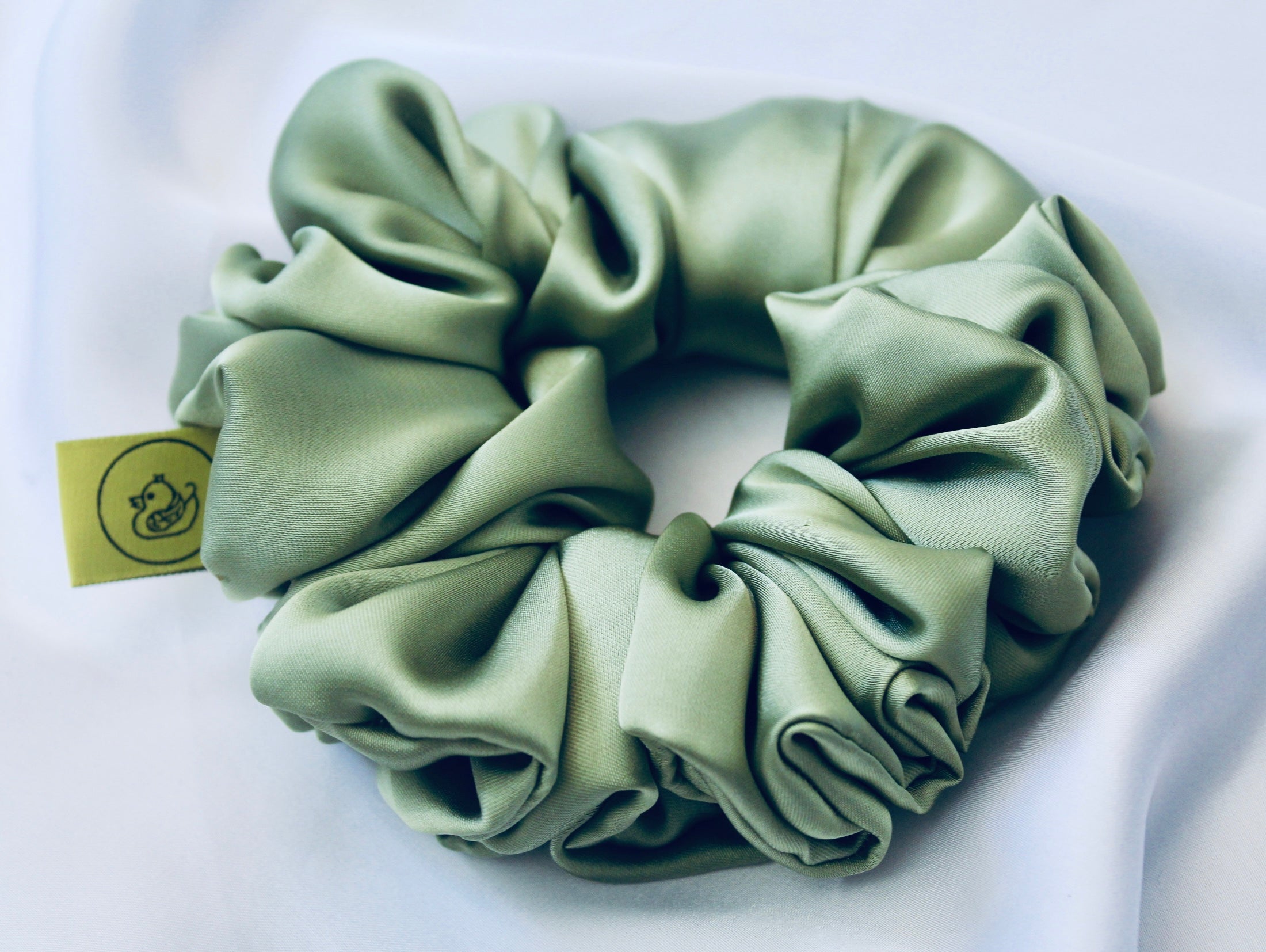 A satin scrunchie in a light green shade with a silky, glossy finish and a ruched design.  Crafted from smooth satin, this scrunchie feels soft and sleek against the hair.  The green tone has a timeless, earthy quality, perfect for both everyday wear and festive styling.  Its ruched fabric is stretched over an elastic band, providing a secure yet comfortable fit.