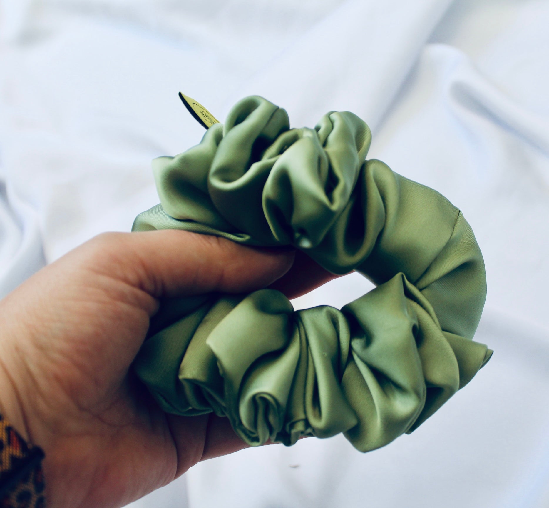 A satin scrunchie in a light green shade with a silky, glossy finish and a ruched design.  Crafted from smooth satin, this scrunchie feels soft and sleek against the hair.  The green tone has a timeless, earthy quality, perfect for both everyday wear and festive styling.  Its ruched fabric is stretched over an elastic band, providing a secure yet comfortable fit.