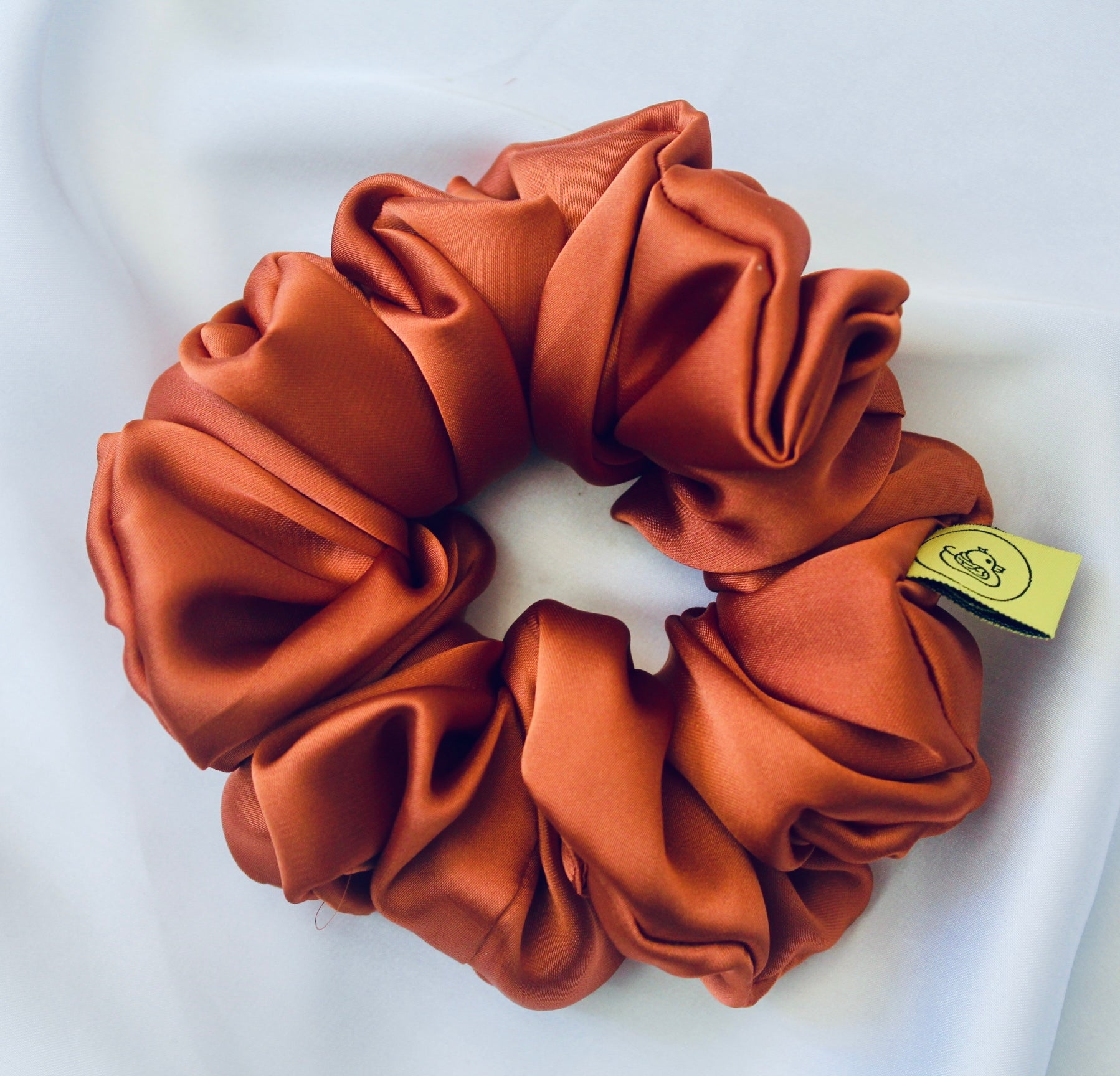 A satin scrunchie in a warm burnt orange shade with a smooth glossy texture and a ruched design.  This scrunchie is made from satin giving it a silky and lightweight feel.  The rich burnt orange colour captures the warmth of autumn leaves, making it perfect seasonal accessory.  The gathered fabric sits on an elastic core, creating a full ruffled look while keeping your hairstyle secure and comfortable.
