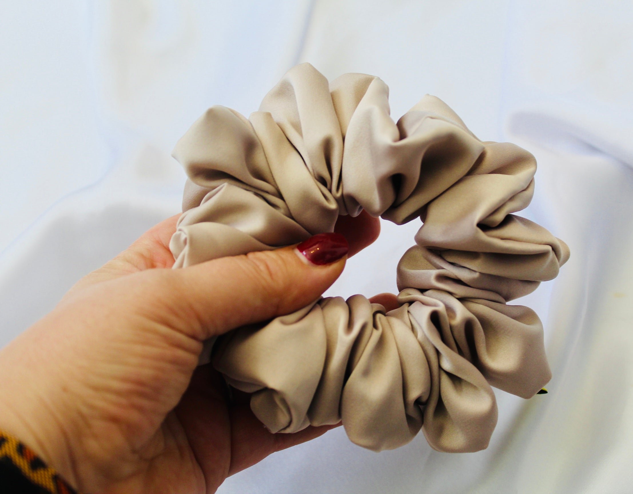 A satin hair scrunchie in a soft taupe beigge with a smooth glossy texture and a ruched design.  This scrunchie is made from satin, giving it a silky, luxurious feel.  The soft taupe shade is a neutral tone that pairs well with any outfit, making it a timeless everyday essential.  It's fabric is gathered along an elastic core, creating a full, ruffled look while staying secure and comfortable in the hair.