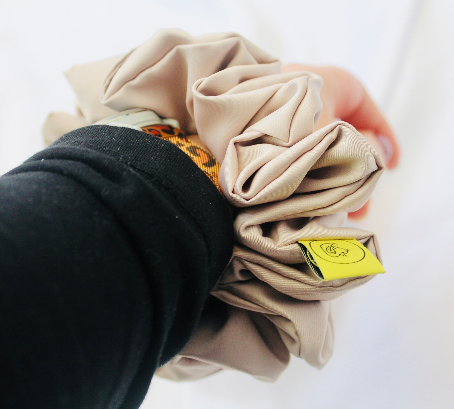 A satin hair scrunchie in a soft taupe beigge with a smooth glossy texture and a ruched design.  This scrunchie is made from satin, giving it a silky, luxurious feel.  The soft taupe shade is a neutral tone that pairs well with any outfit, making it a timeless everyday essential.  It's fabric is gathered along an elastic core, creating a full, ruffled look while staying secure and comfortable in the hair.