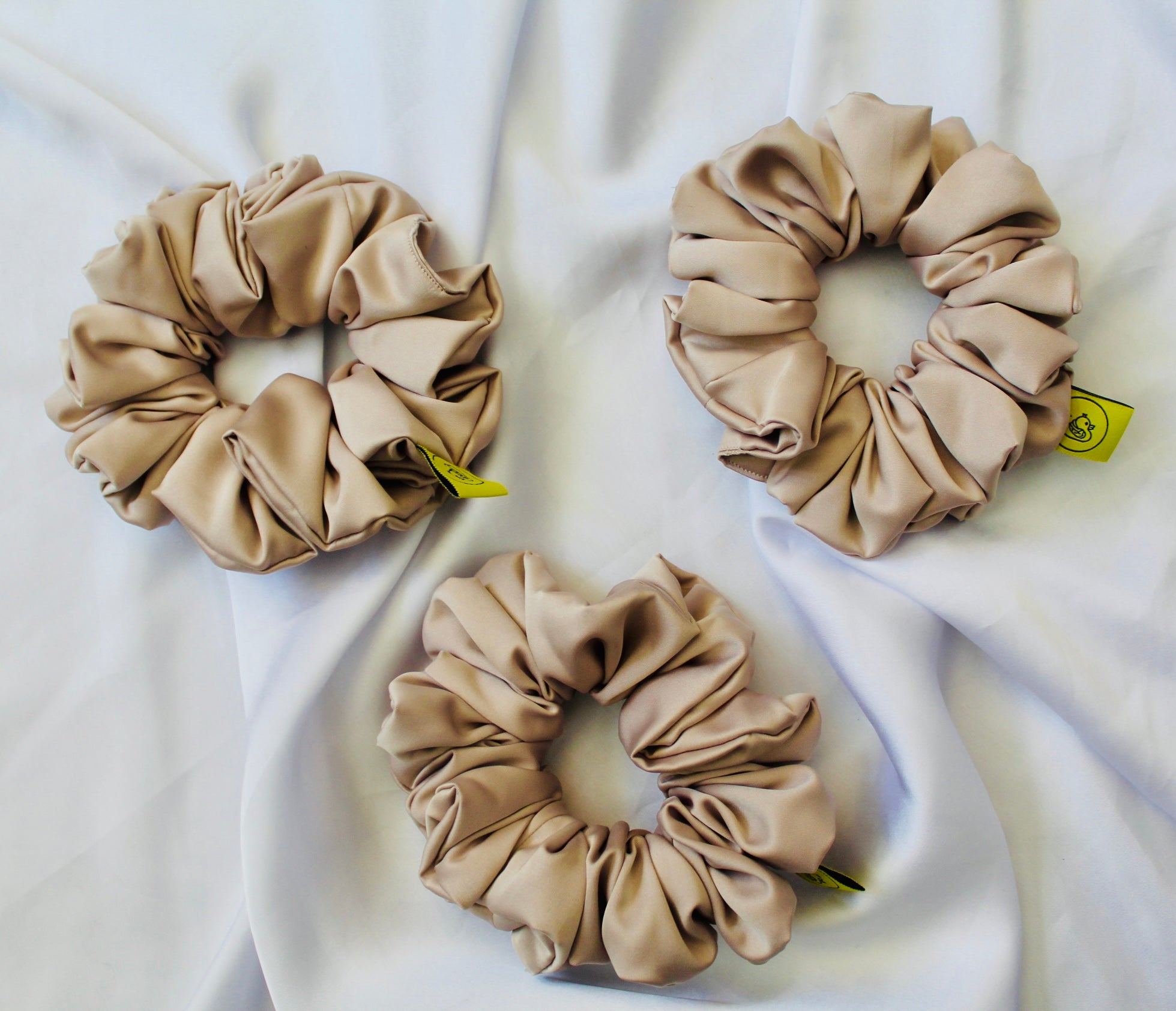 A satin hair scrunchie in a soft taupe beigge with a smooth glossy texture and a ruched design.  This scrunchie is made from satin, giving it a silky, luxurious feel.  The soft taupe shade is a neutral tone that pairs well with any outfit, making it a timeless everyday essential.  It's fabric is gathered along an elastic core, creating a full, ruffled look while staying secure and comfortable in the hair.