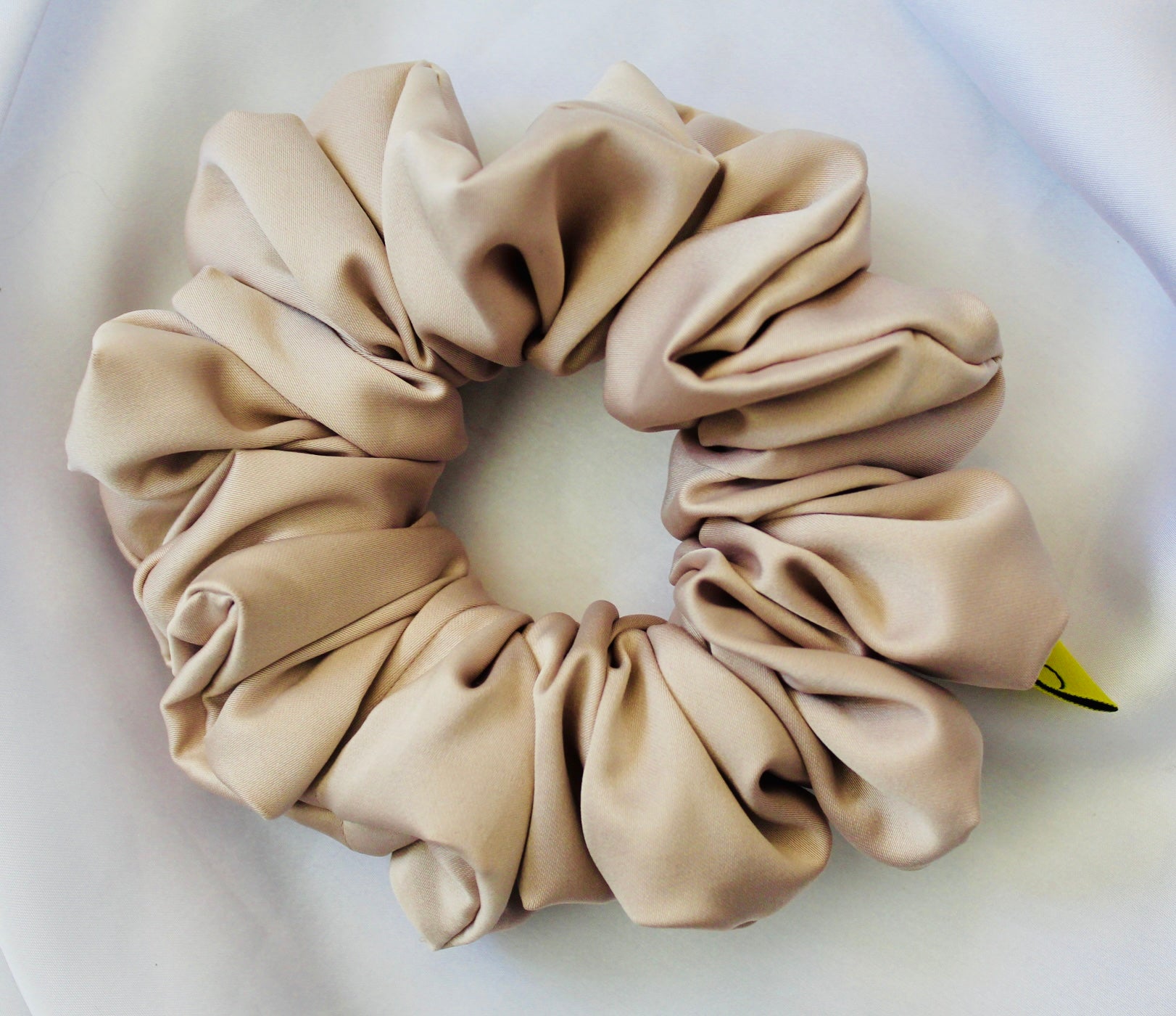 A satin hair scrunchie in a soft taupe beigge with a smooth glossy texture and a ruched design.  This scrunchie is made from satin, giving it a silky, luxurious feel.  The soft taupe shade is a neutral tone that pairs well with any outfit, making it a timeless everyday essential.  It's fabric is gathered along an elastic core, creating a full, ruffled look while staying secure and comfortable in the hair.