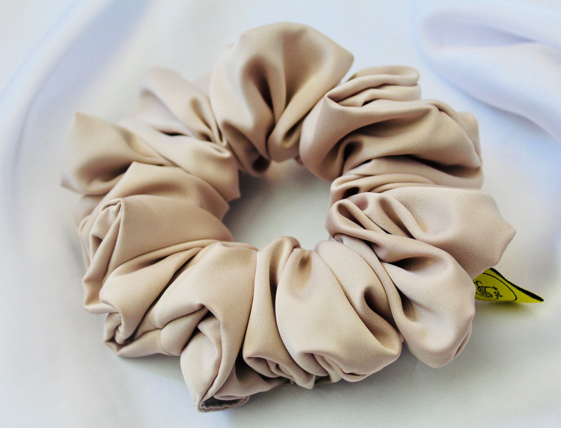 A satin hair scrunchie in a soft taupe beigge with a smooth glossy texture and a ruched design.  This scrunchie is made from satin, giving it a silky, luxurious feel.  The soft taupe shade is a neutral tone that pairs well with any outfit, making it a timeless everyday essential.  It's fabric is gathered along an elastic core, creating a full, ruffled look while staying secure and comfortable in the hair.