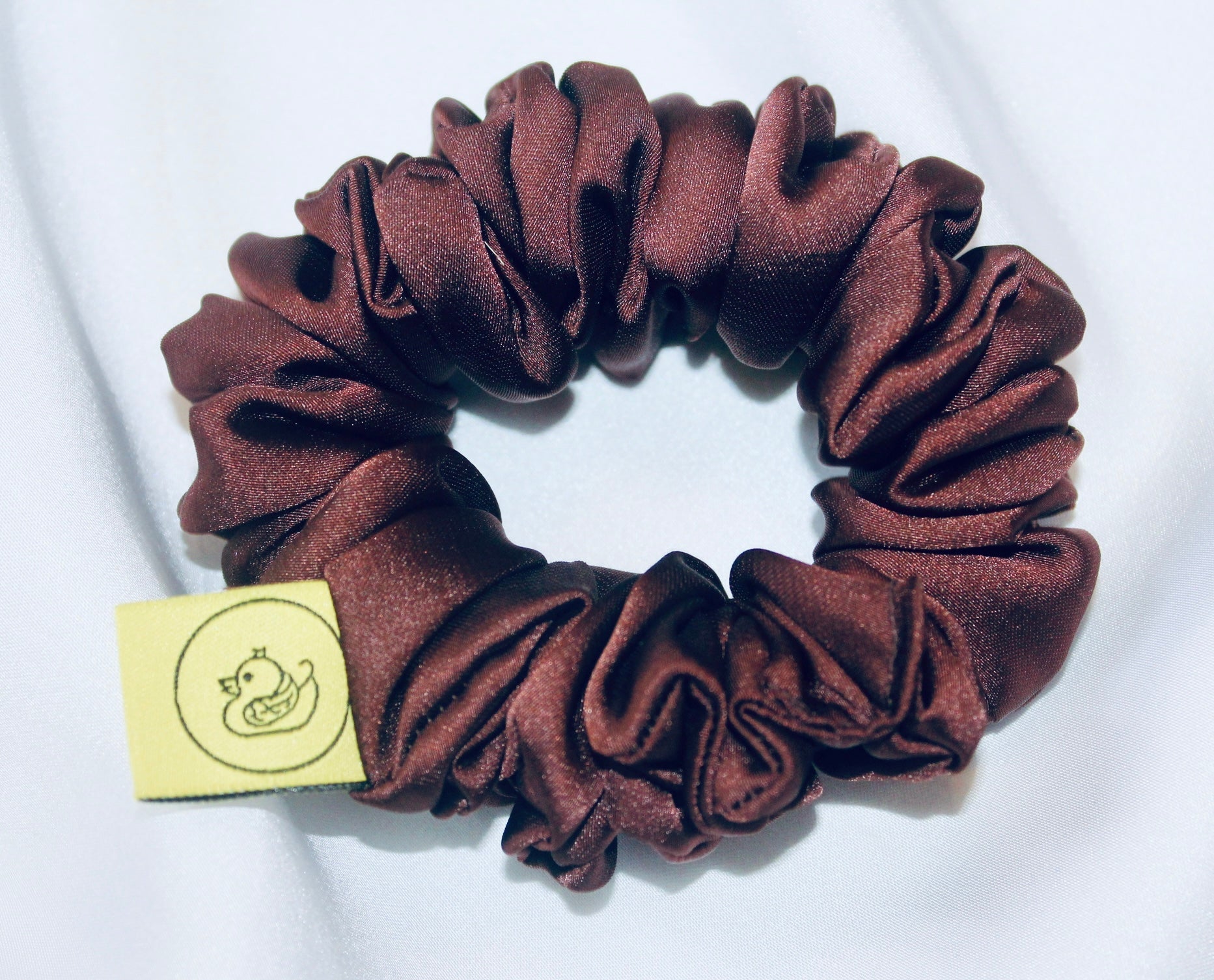 A satin scrunchie in a rich chocolate brown shade with a smooth and shiny texture and a ruched design.  This scrunchie is made from glossy satin giving it a soft, luxurious touch.  The deep chocolate brown colour is both bold and neutral, making it easy to style with a wide range of outfits.  The fabric is softly ruched over elastic, creating volume while remaining comfortable to wear.