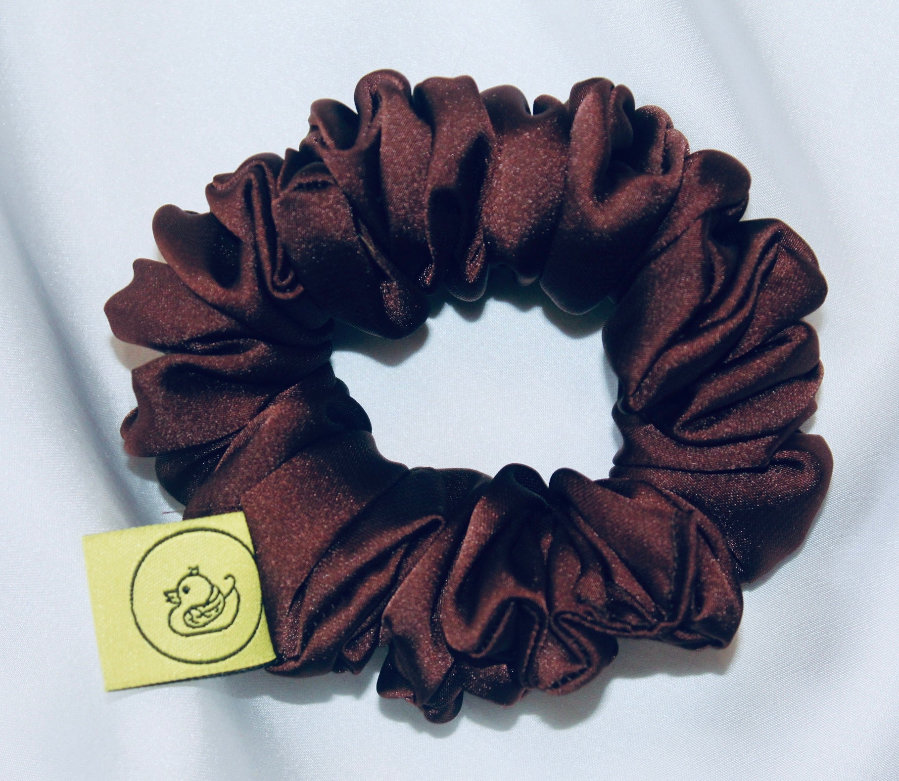 A satin scrunchie in a rich chocolate brown shade with a smooth and shiny texture and a ruched design.  This scrunchie is made from glossy satin giving it a soft, luxurious touch.  The deep chocolate brown colour is both bold and neutral, making it easy to style with a wide range of outfits.  The fabric is softly ruched over elastic, creating volume while remaining comfortable to wear.