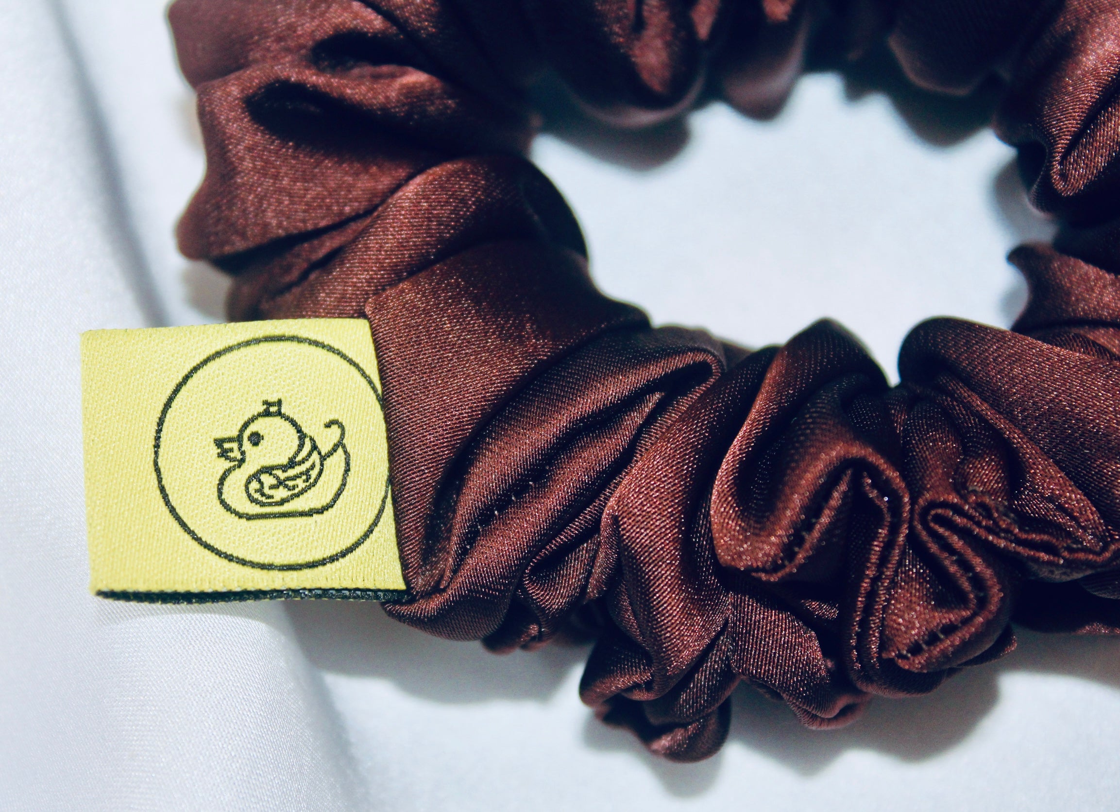A satin scrunchie in a rich chocolate brown shade with a smooth and shiny texture and a ruched design.  This scrunchie is made from glossy satin giving it a soft, luxurious touch.  The deep chocolate brown colour is both bold and neutral, making it easy to style with a wide range of outfits.  The fabric is softly ruched over elastic, creating volume while remaining comfortable to wear.