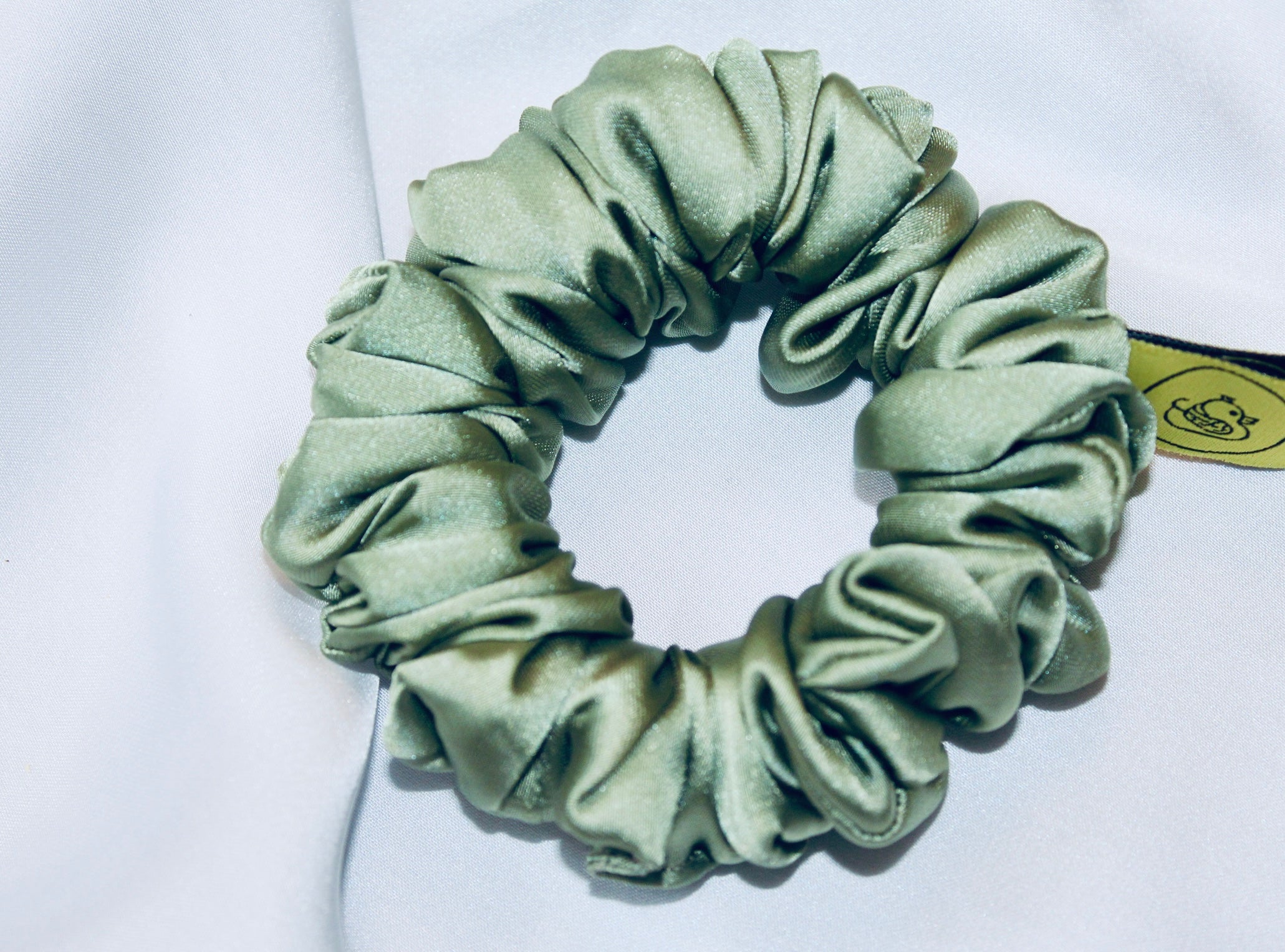 A satin scrunchie in a light green shade with a silky, glossy finish and a ruched design.  Crafted from smooth satin, this scrunchie feels soft and sleek against the hair.  The green tone has a timeless, earthy quality, perfect for both everyday wear and festive styling.  Its ruched fabric is stretched over an elastic band, providing a secure yet comfortable fit.