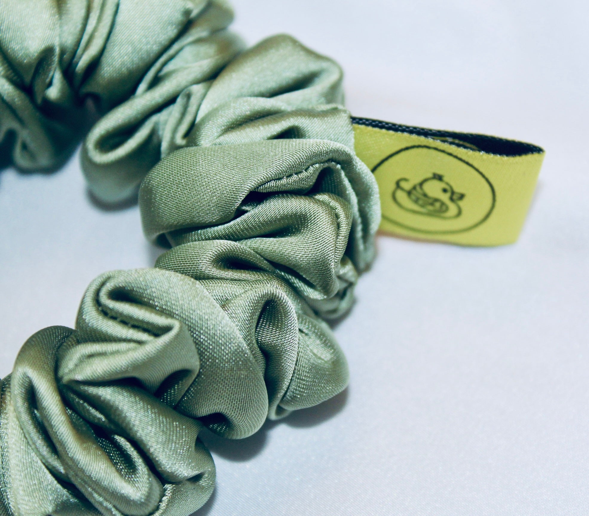 A satin scrunchie in a light green shade with a silky, glossy finish and a ruched design.  Crafted from smooth satin, this scrunchie feels soft and sleek against the hair.  The green tone has a timeless, earthy quality, perfect for both everyday wear and festive styling.  Its ruched fabric is stretched over an elastic band, providing a secure yet comfortable fit.