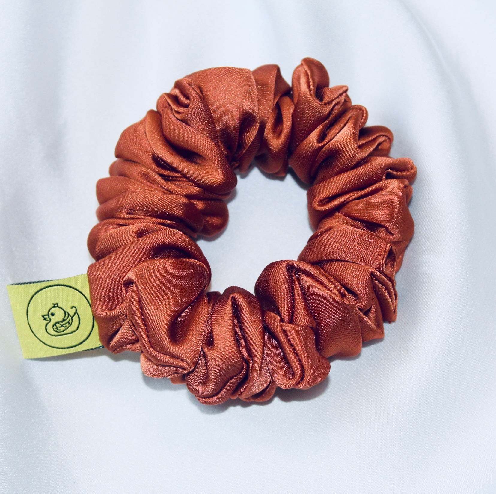 A satin scrunchie in a warm burnt orange shade with a smooth glossy texture and a ruched design.  This scrunchie is made from satin giving it a silky and lightweight feel.  The rich burnt orange colour captures the warmth of autumn leaves, making it perfect seasonal accessory.  The gathered fabric sits on an elastic core, creating a full ruffled look while keeping your hairstyle secure and comfortable.