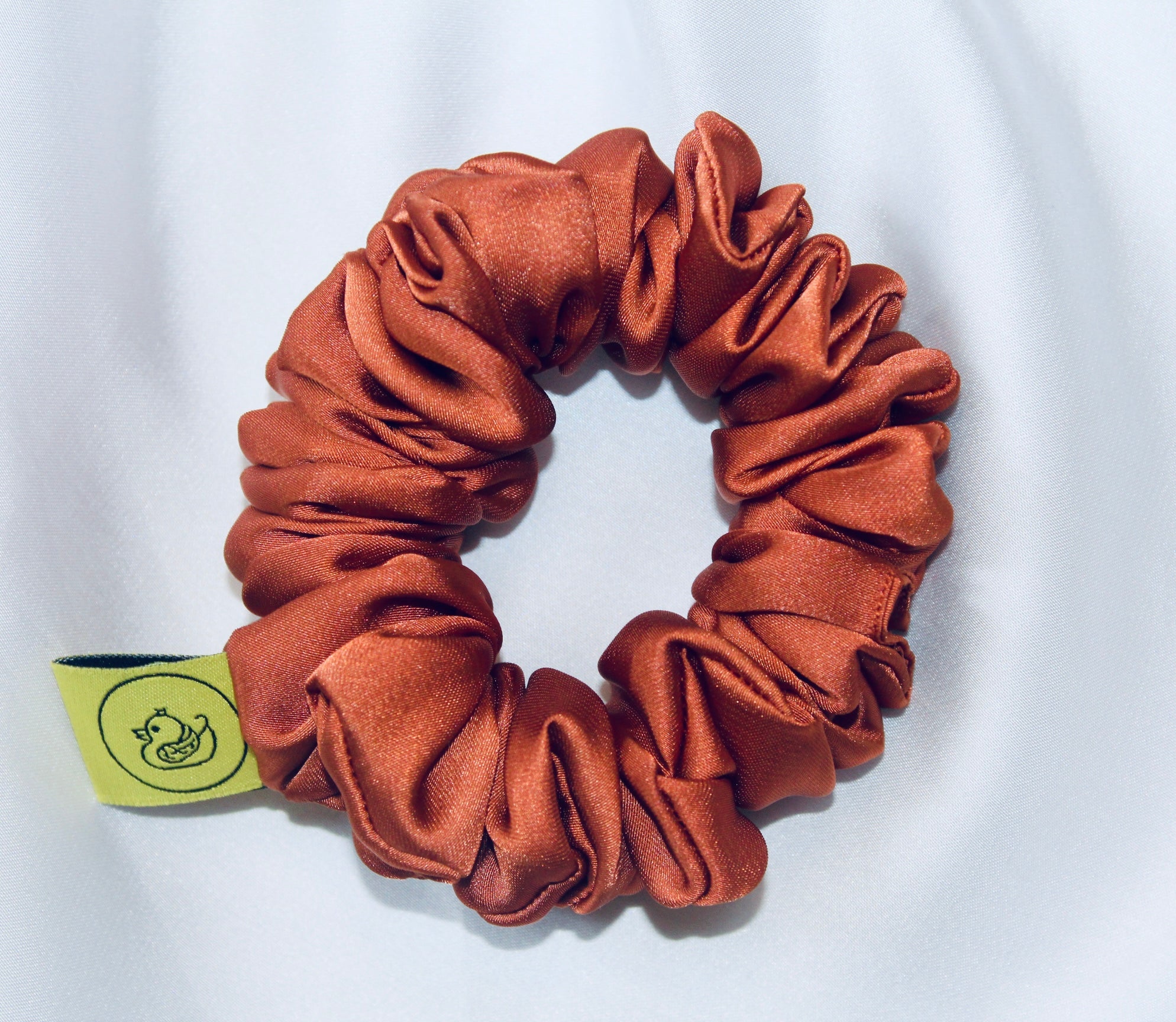 A satin scrunchie in a warm burnt orange shade with a smooth glossy texture and a ruched design.  This scrunchie is made from satin giving it a silky and lightweight feel.  The rich burnt orange colour captures the warmth of autumn leaves, making it perfect seasonal accessory.  The gathered fabric sits on an elastic core, creating a full ruffled look while keeping your hairstyle secure and comfortable.