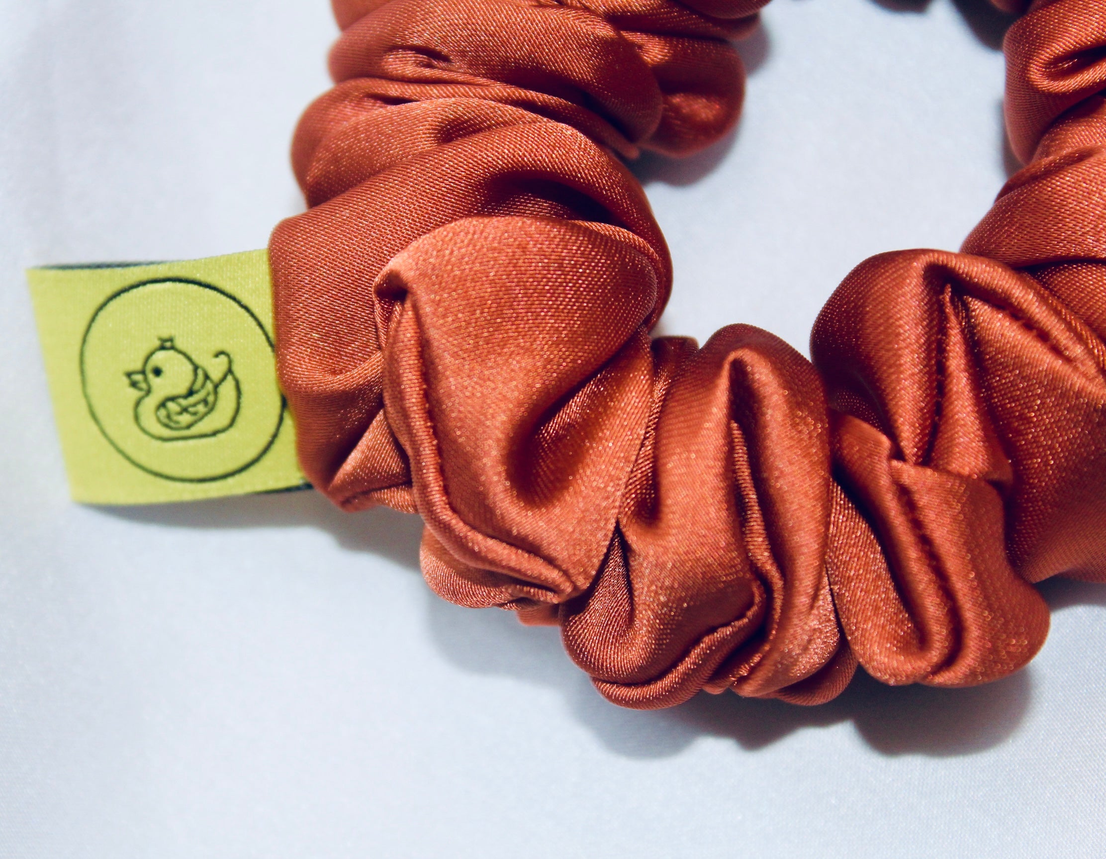 A satin scrunchie in a warm burnt orange shade with a smooth glossy texture and a ruched design.  This scrunchie is made from satin giving it a silky and lightweight feel.  The rich burnt orange colour captures the warmth of autumn leaves, making it perfect seasonal accessory.  The gathered fabric sits on an elastic core, creating a full ruffled look while keeping your hairstyle secure and comfortable.