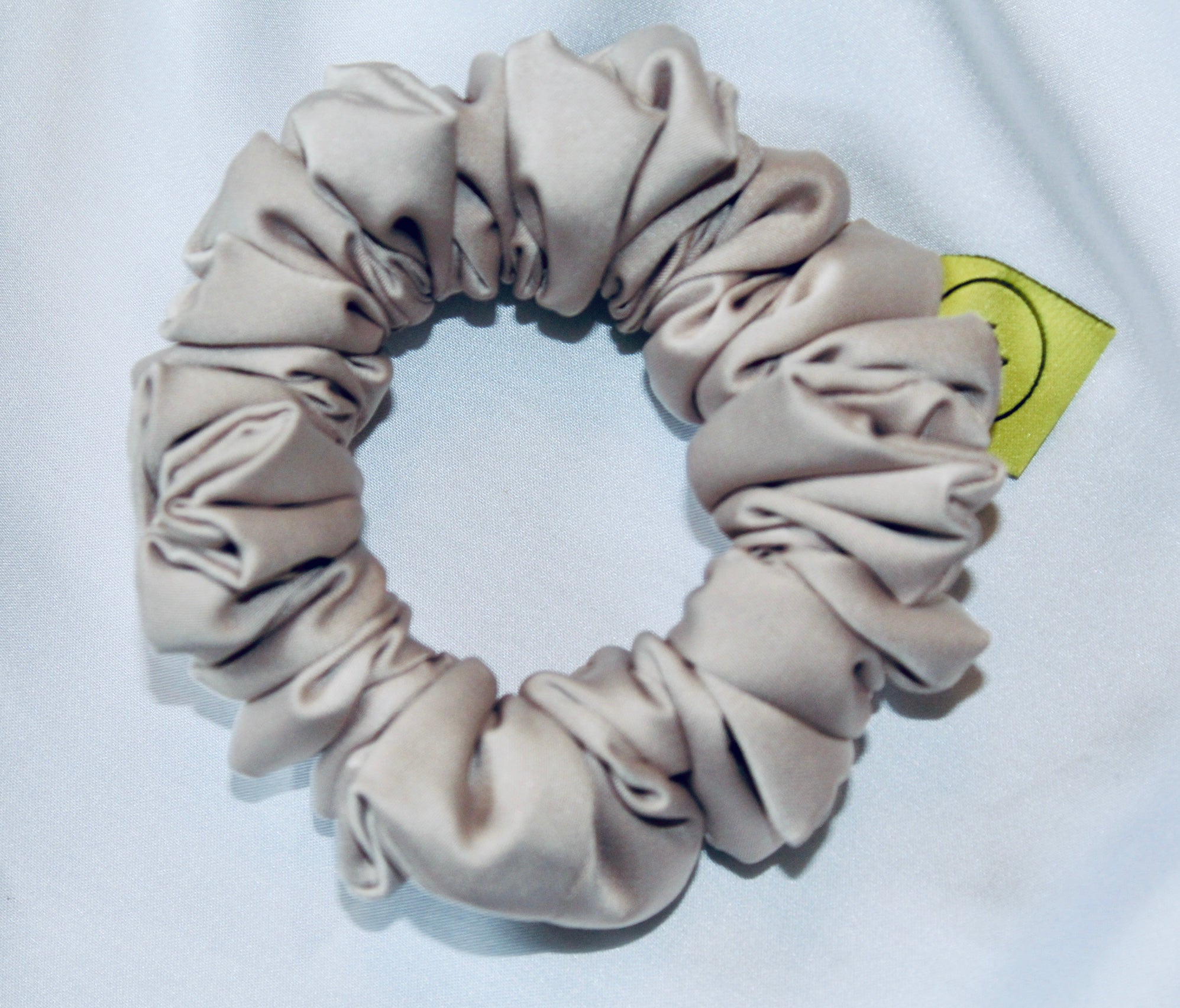 A satin hair scrunchie in a soft taupe beigge with a smooth glossy texture and a ruched design.  This scrunchie is made from satin, giving it a silky, luxurious feel.  The soft taupe shade is a neutral tone that pairs well with any outfit, making it a timeless everyday essential.  It's fabric is gathered along an elastic core, creating a full, ruffled look while staying secure and comfortable in the hair.