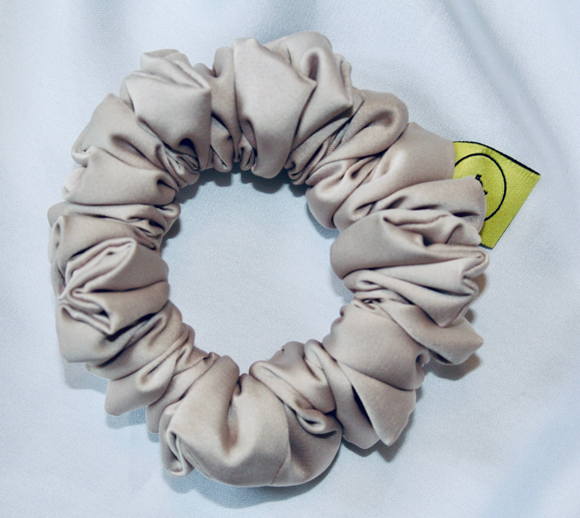 A satin hair scrunchie in a soft taupe beigge with a smooth glossy texture and a ruched design.  This scrunchie is made from satin, giving it a silky, luxurious feel.  The soft taupe shade is a neutral tone that pairs well with any outfit, making it a timeless everyday essential.  It's fabric is gathered along an elastic core, creating a full, ruffled look while staying secure and comfortable in the hair.