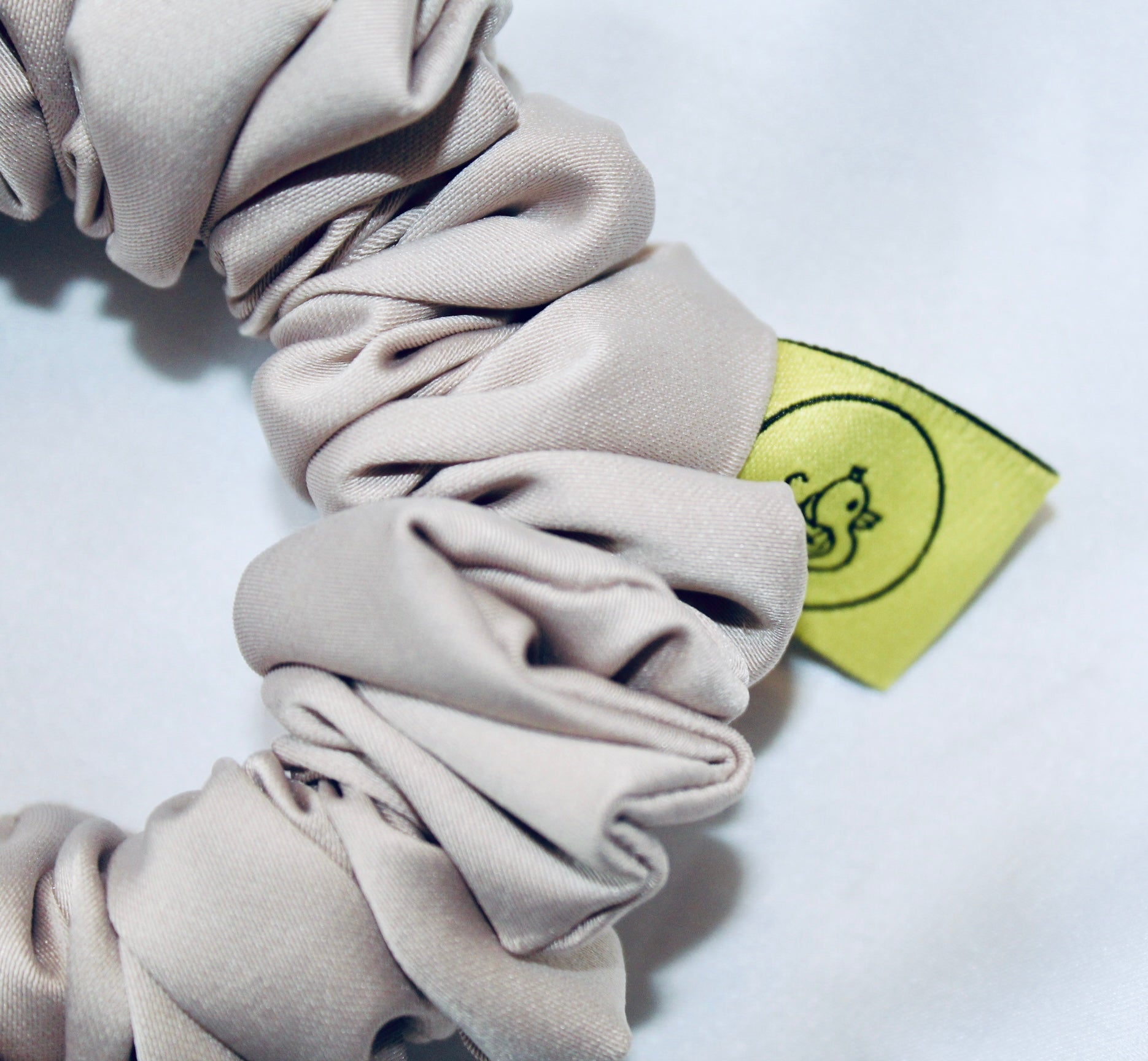 A satin hair scrunchie in a soft taupe beigge with a smooth glossy texture and a ruched design.  This scrunchie is made from satin, giving it a silky, luxurious feel.  The soft taupe shade is a neutral tone that pairs well with any outfit, making it a timeless everyday essential.  It's fabric is gathered along an elastic core, creating a full, ruffled look while staying secure and comfortable in the hair.