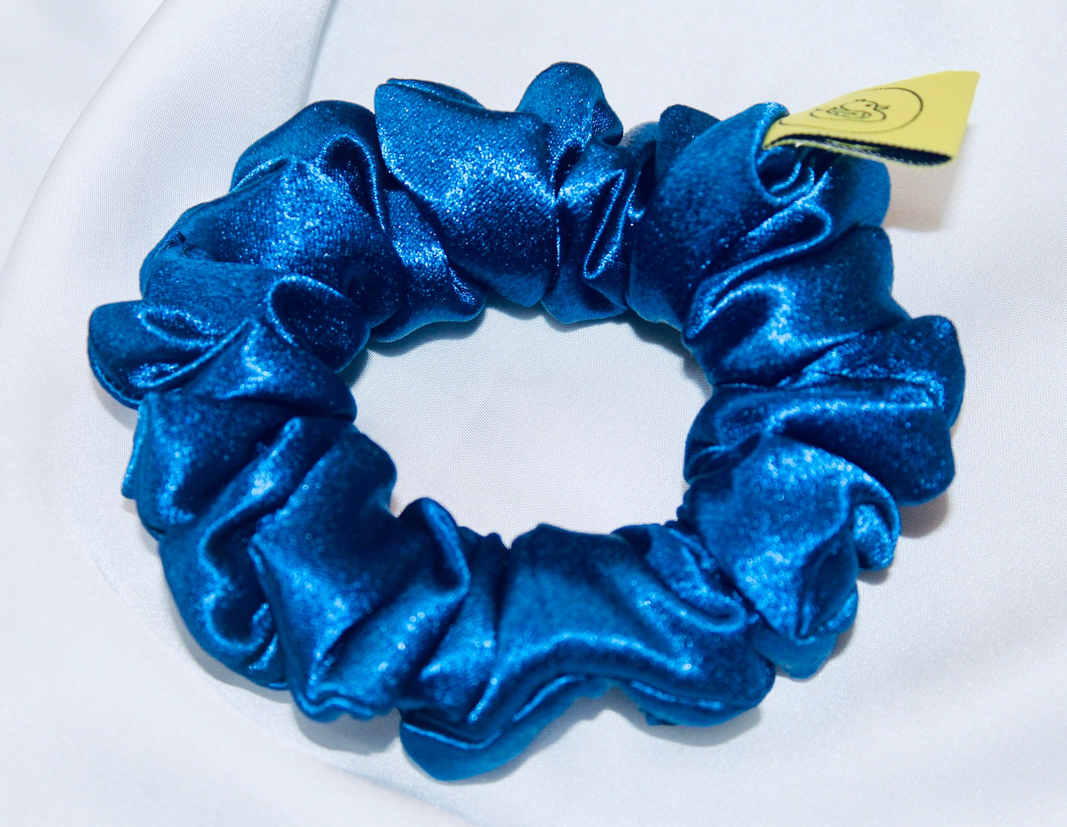 A satin scrunchie in a striking royal blue shade with a silky glossy texture and a ruched design.  Crafted from satin this scrunchie feels smooth and cool to the touch.  The bold royal blue colour stands out beautifully, making it a statement piece for both casual and evening looks.  The gathered satin fabric creates a full textured effect over the elastic ensuring a secure hold.