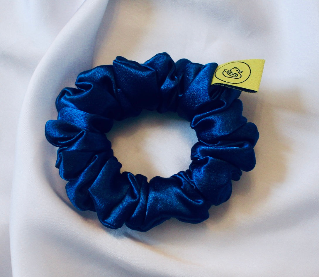A satin scrunchie in a striking royal blue shade with a silky glossy texture and a ruched design.  Crafted from satin this scrunchie feels smooth and cool to the touch.  The bold royal blue colour stands out beautifully, making it a statement piece for both casual and evening looks.  The gathered satin fabric creates a full textured effect over the elastic ensuring a secure hold.