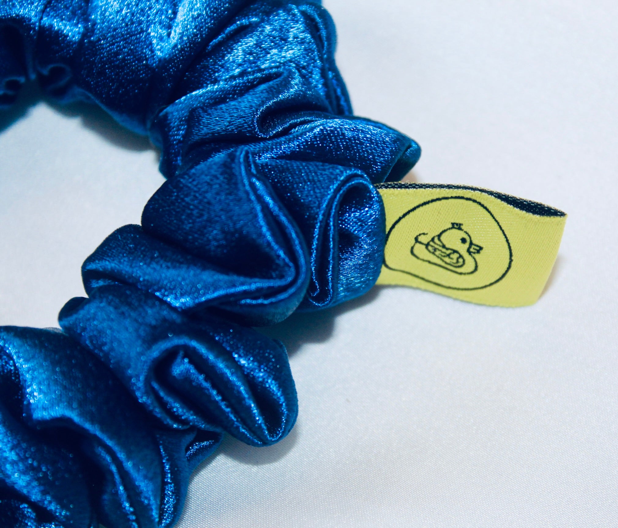 A satin scrunchie in a striking royal blue shade with a silky glossy texture and a ruched design.  Crafted from satin this scrunchie feels smooth and cool to the touch.  The bold royal blue colour stands out beautifully, making it a statement piece for both casual and evening looks.  The gathered satin fabric creates a full textured effect over the elastic ensuring a secure hold.