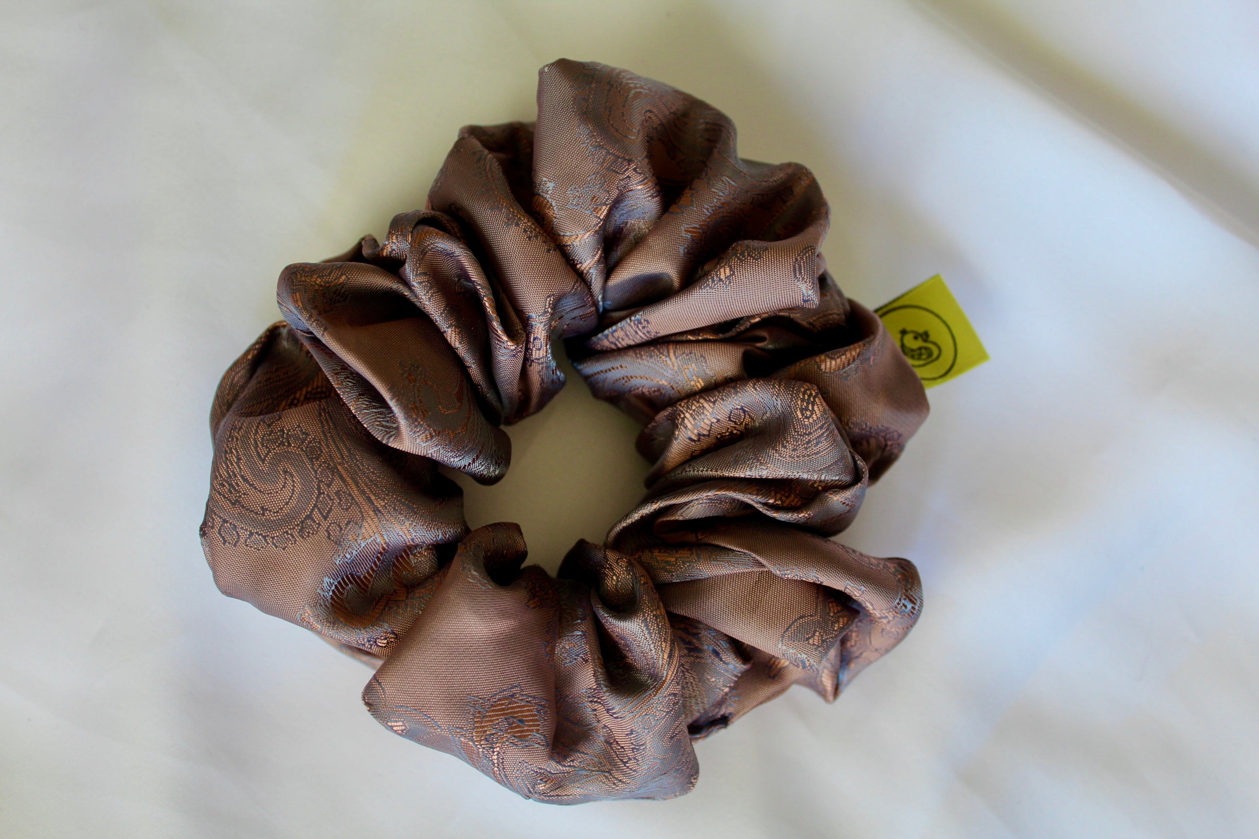 This shows a luxurious scrunchie made of silky textured fabric in a brown mocha and gold.The fabric has a subtle paisley pattern wove into it, giving it an elegant and slightly reflective appearance.The scrunchie is gathered and voluminous creating soft folds and a plush look.A small yellow label tag with a picture of a duck is attached to the scrunchie adding a touch of branding.The overall aesthetic is soft, elegant and stylish. Perfect for those with sensitive scalp or hair loss