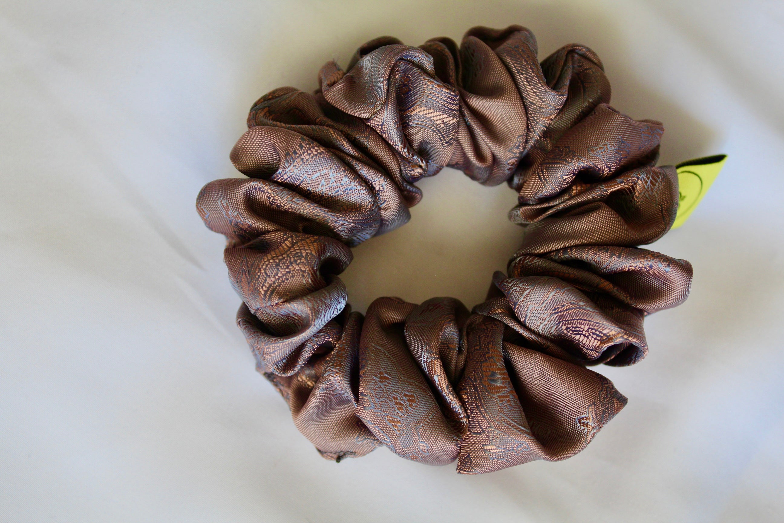 This shows a luxurious scrunchie made of silky textured fabric in a brown mocha and gold.The fabric has a subtle paisley pattern wove into it, giving it an elegant and slightly reflective appearance.The scrunchie is gathered and voluminous creating soft folds and a plush look.A small yellow label tag with a picture of a duck is attached to the scrunchie adding a touch of branding.The overall aesthetic is soft, elegant and stylish. Perfect for those with sensitive scalp or hair loss