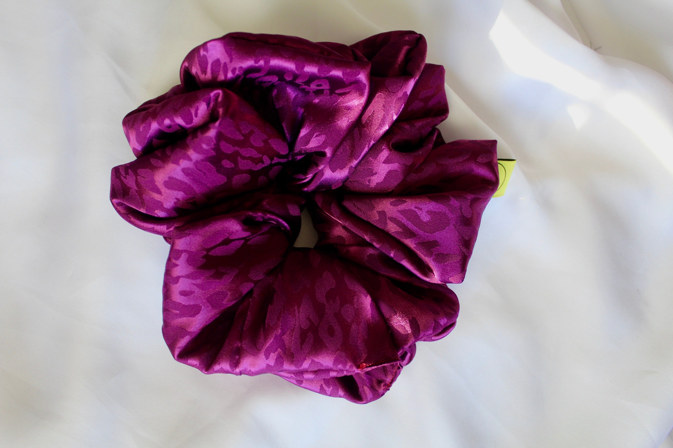 This shows a luxurious scrunchie made of silky textured fabric in a bright deep purple colour.The fabric has a subtle leopard print pattern wove into it, giving it an elegant and slightly reflective appearance.The scrunchie is gathered and voluminous creating soft folds and a plush look.A small yellow label tag with a picture of a duck is attached to the scrunchie adding a touch of branding.The overall aesthetic is soft, elegant and stylish. Perfect for those with sensitive scalp or hair loss