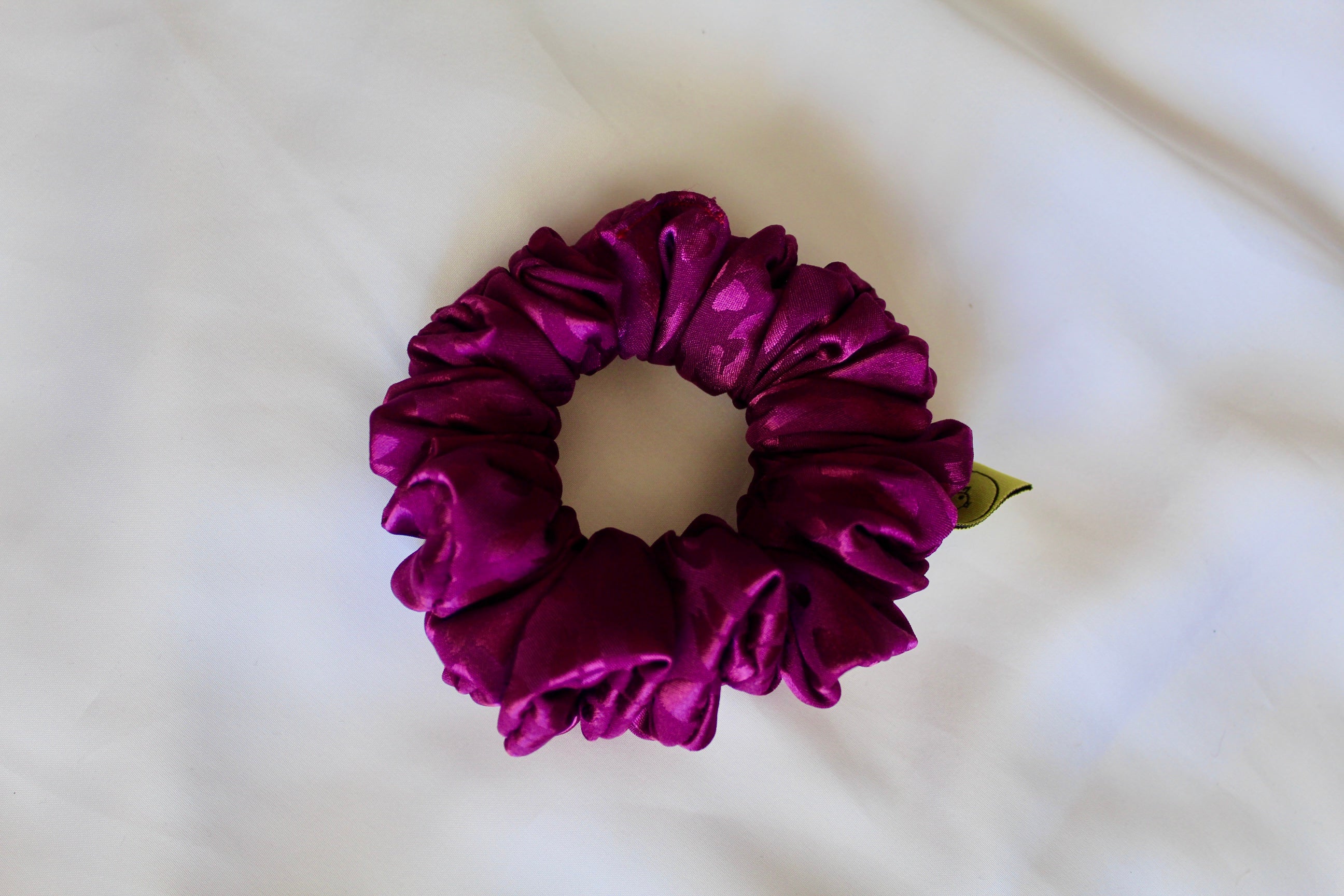 This shows a luxurious scrunchie made of silky textured fabric in a bright deep purple colour.The fabric has a subtle leopard print pattern wove into it, giving it an elegant and slightly reflective appearance.The scrunchie is gathered and voluminous creating soft folds and a plush look.A small yellow label tag with a picture of a duck is attached to the scrunchie adding a touch of branding.The overall aesthetic is soft, elegant and stylish. Perfect for those with sensitive scalp or hair loss