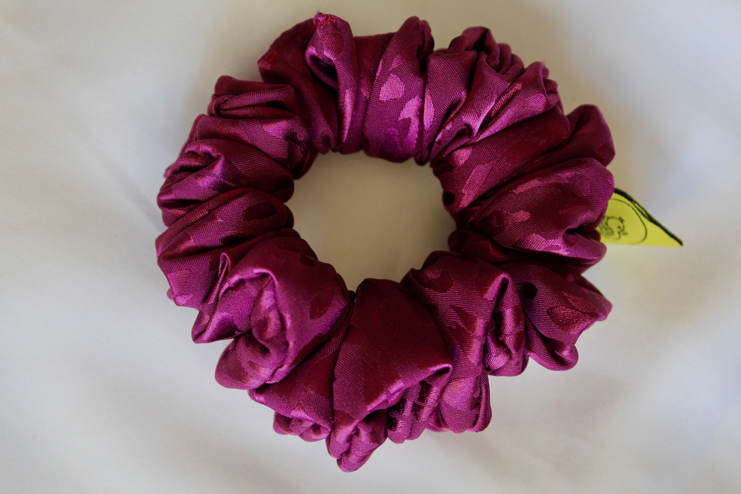 This shows a luxurious scrunchie made of silky textured fabric in a bright deep purple colour.The fabric has a subtle leopard print pattern wove into it, giving it an elegant and slightly reflective appearance.The scrunchie is gathered and voluminous creating soft folds and a plush look.A small yellow label tag with a picture of a duck is attached to the scrunchie adding a touch of branding.The overall aesthetic is soft, elegant and stylish. Perfect for those with sensitive scalp or hair loss