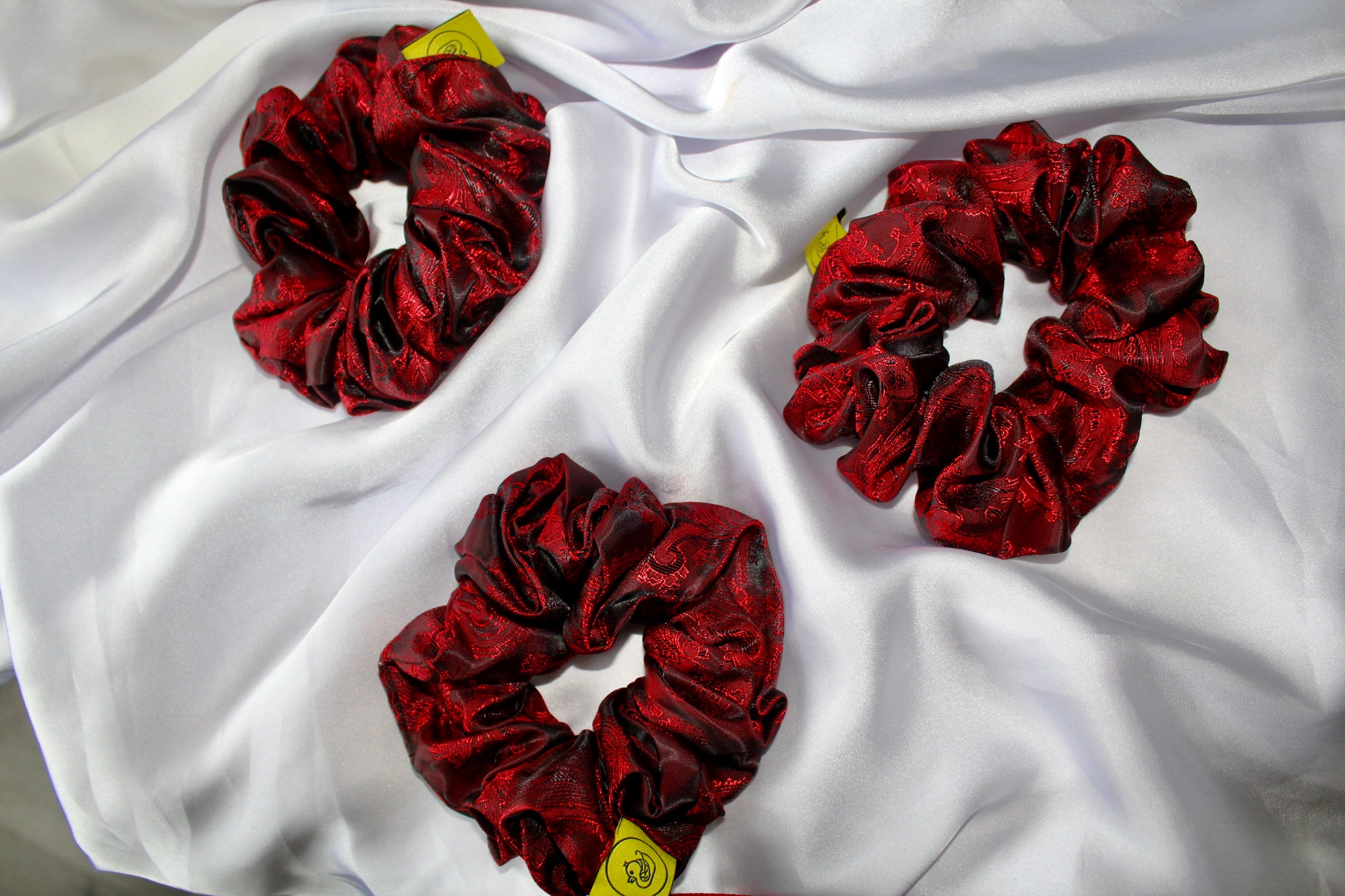 This shows a luxurious scrunchie made of silky textured fabric in a red and black colour.The fabric has a subtle paisley pattern wove into it, giving it an elegant and slightly reflective appearance.The scrunchie is gathered and voluminous creating soft folds and a plush look.A small yellow label tag with a picture of a duck is attached to the scrunchie adding a touch of branding.The overall aesthetic is soft, elegant and stylish. Perfect for those with sensitive scalp or hair loss