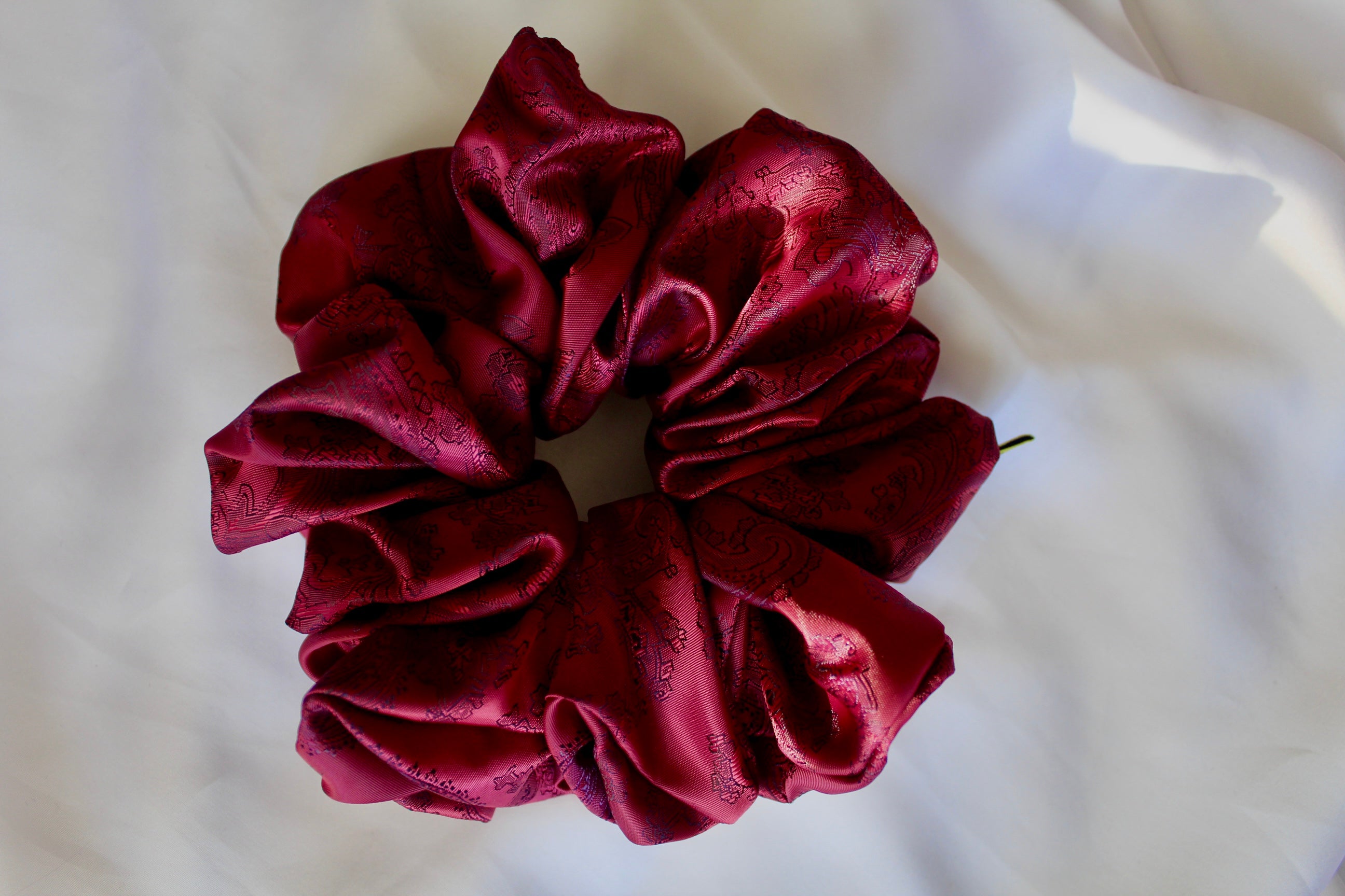 This shows a luxurious scrunchie made of silky textured fabric in red and blue paisley patter running through the fabric giving it an elegant and slightly reflective appearance.The scrunchie is gathered and voluminous creating soft folds and a plush look.A small yellow label tag with a picture of a duck is attached to the scrunchie adding a touch of branding.The overall aesthetic is soft, elegant and stylish. Perfect for those with sensitive scalp or hair loss