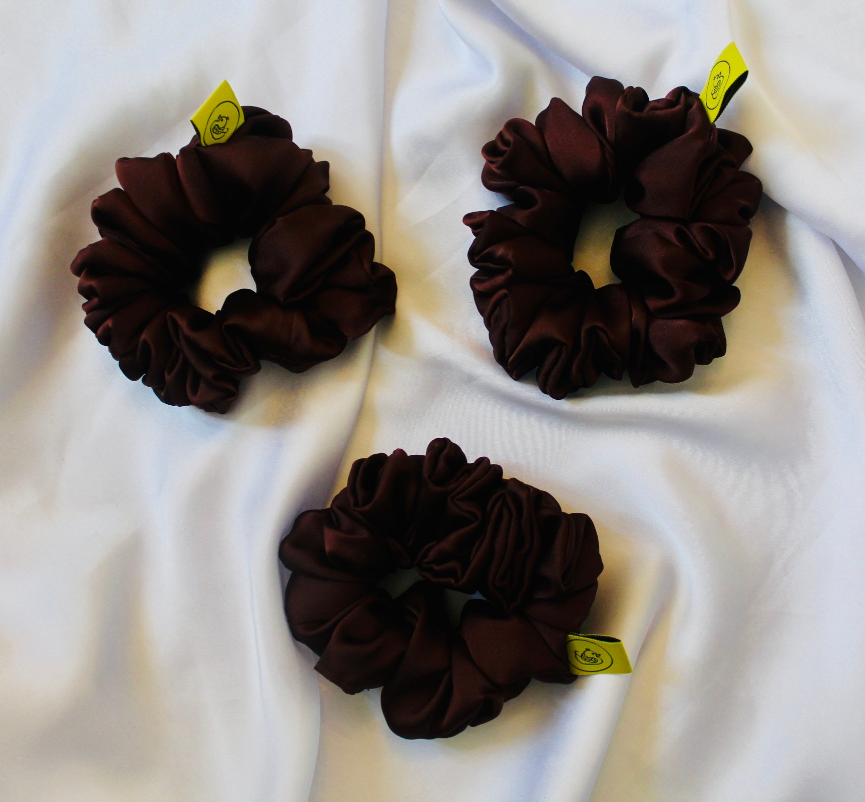 A satin scrunchie in a rich chocolate brown shade with a smooth and shiny texture and a ruched design.  This scrunchie is made from glossy satin giving it a soft, luxurious touch.  The deep chocolate brown colour is both bold and neutral, making it easy to style with a wide range of outfits.  The fabric is softly ruched over elastic, creating volume while remaining comfortable to wear.