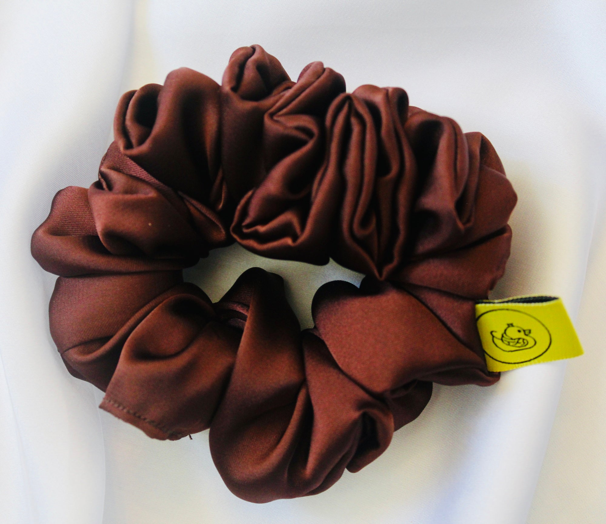 A satin scrunchie in a rich chocolate brown shade with a smooth and shiny texture and a ruched design.  This scrunchie is made from glossy satin giving it a soft, luxurious touch.  The deep chocolate brown colour is both bold and neutral, making it easy to style with a wide range of outfits.  The fabric is softly ruched over elastic, creating volume while remaining comfortable to wear.