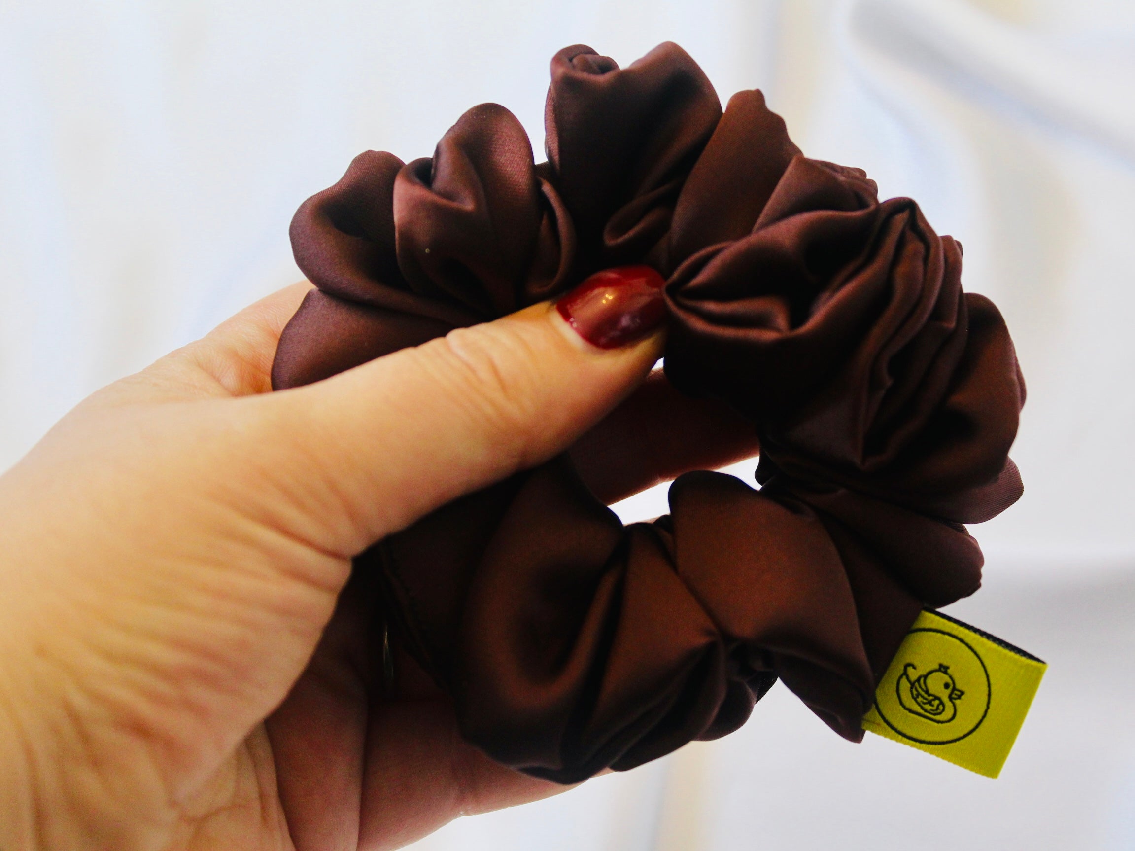 A satin scrunchie in a rich chocolate brown shade with a smooth and shiny texture and a ruched design.  This scrunchie is made from glossy satin giving it a soft, luxurious touch.  The deep chocolate brown colour is both bold and neutral, making it easy to style with a wide range of outfits.  The fabric is softly ruched over elastic, creating volume while remaining comfortable to wear.