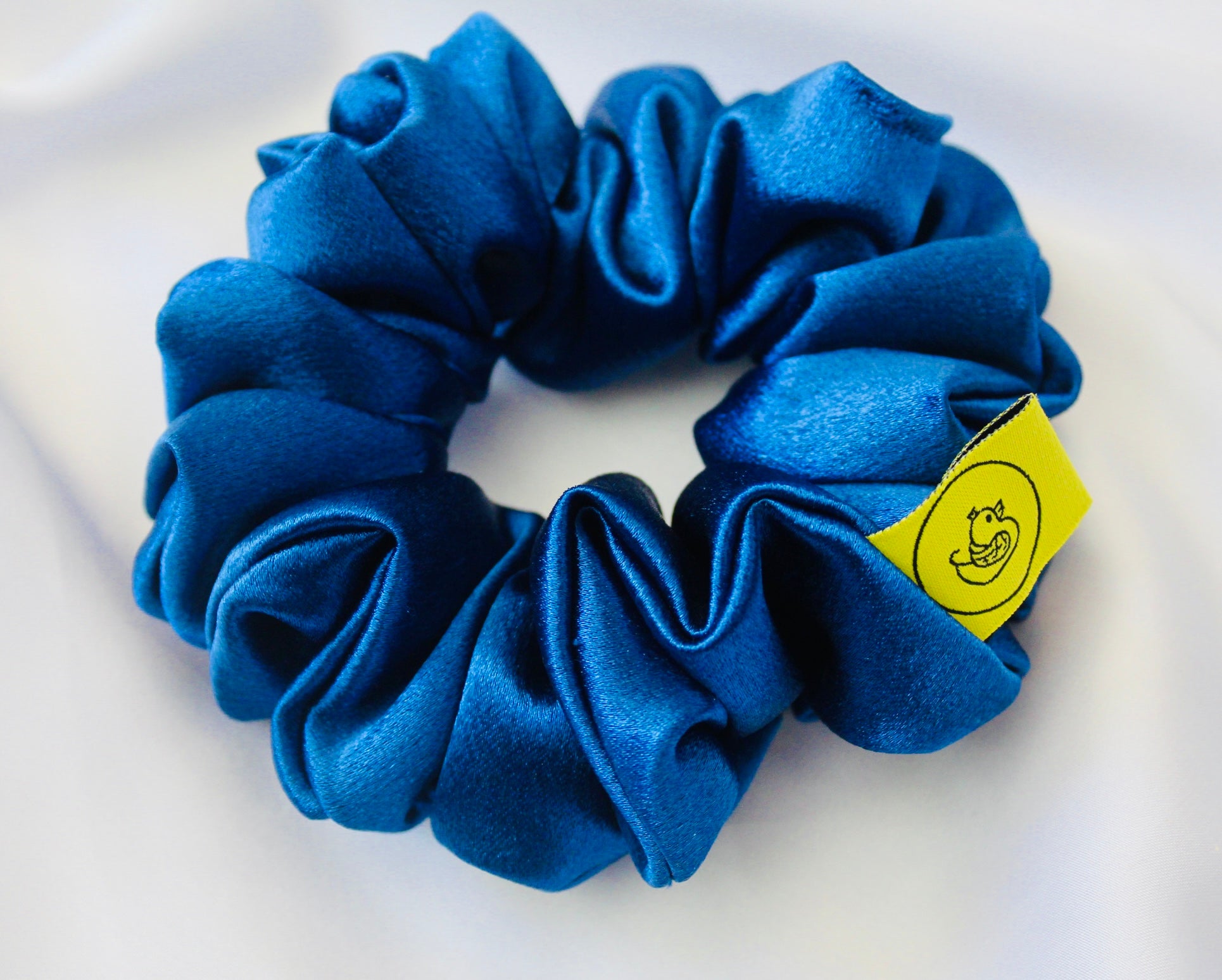 A satin scrunchie in a striking royal blue shade with a silky glossy texture and a ruched design.  Crafted from satin this scrunchie feels smooth and cool to the touch.  The bold royal blue colour stands out beautifully, making it a statement piece for both casual and evening looks.  The gathered satin fabric creates a full textured effect over the elastic ensuring a secure hold.