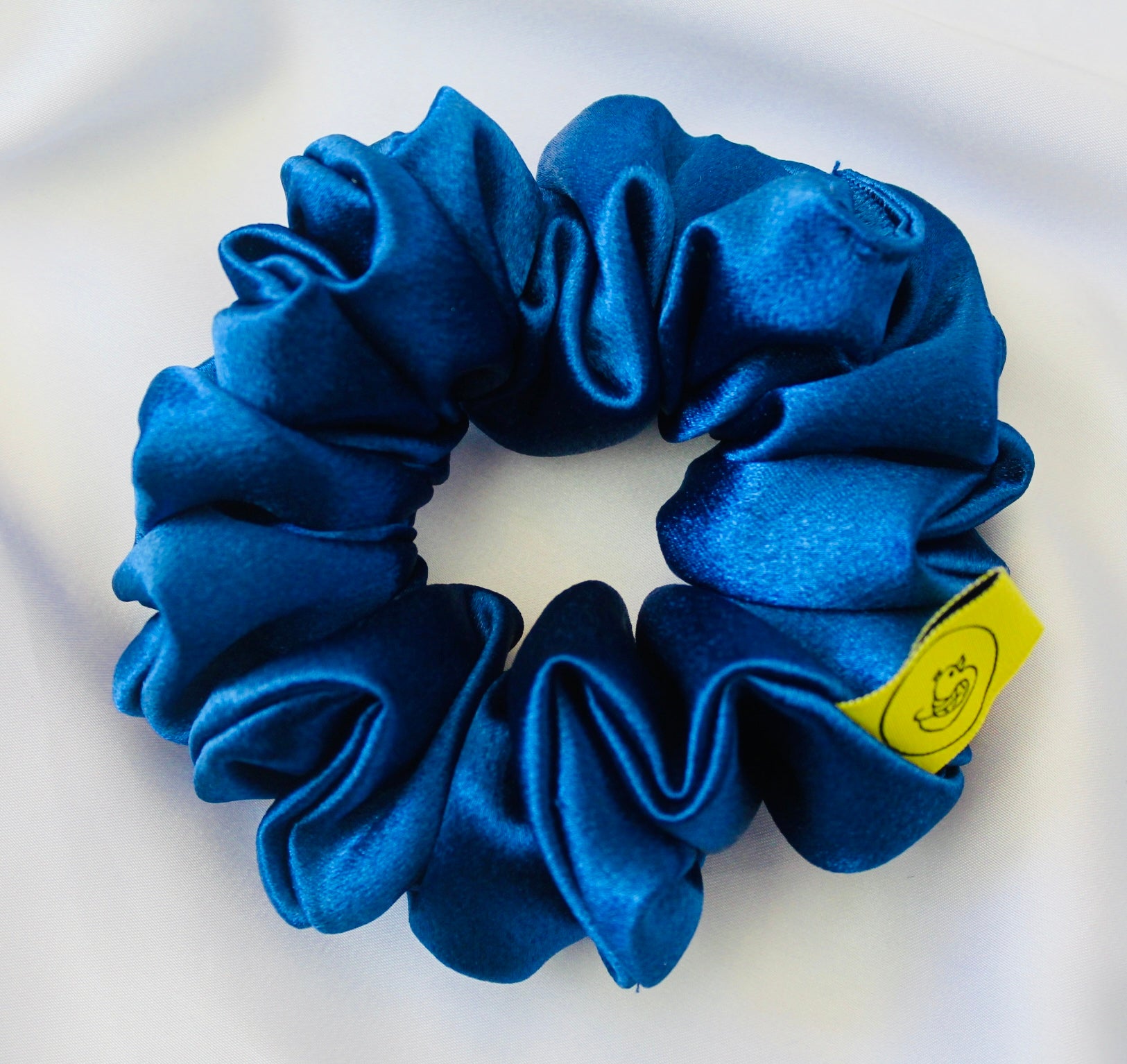 A satin scrunchie in a striking royal blue shade with a silky glossy texture and a ruched design.  Crafted from satin this scrunchie feels smooth and cool to the touch.  The bold royal blue colour stands out beautifully, making it a statement piece for both casual and evening looks.  The gathered satin fabric creates a full textured effect over the elastic ensuring a secure hold.