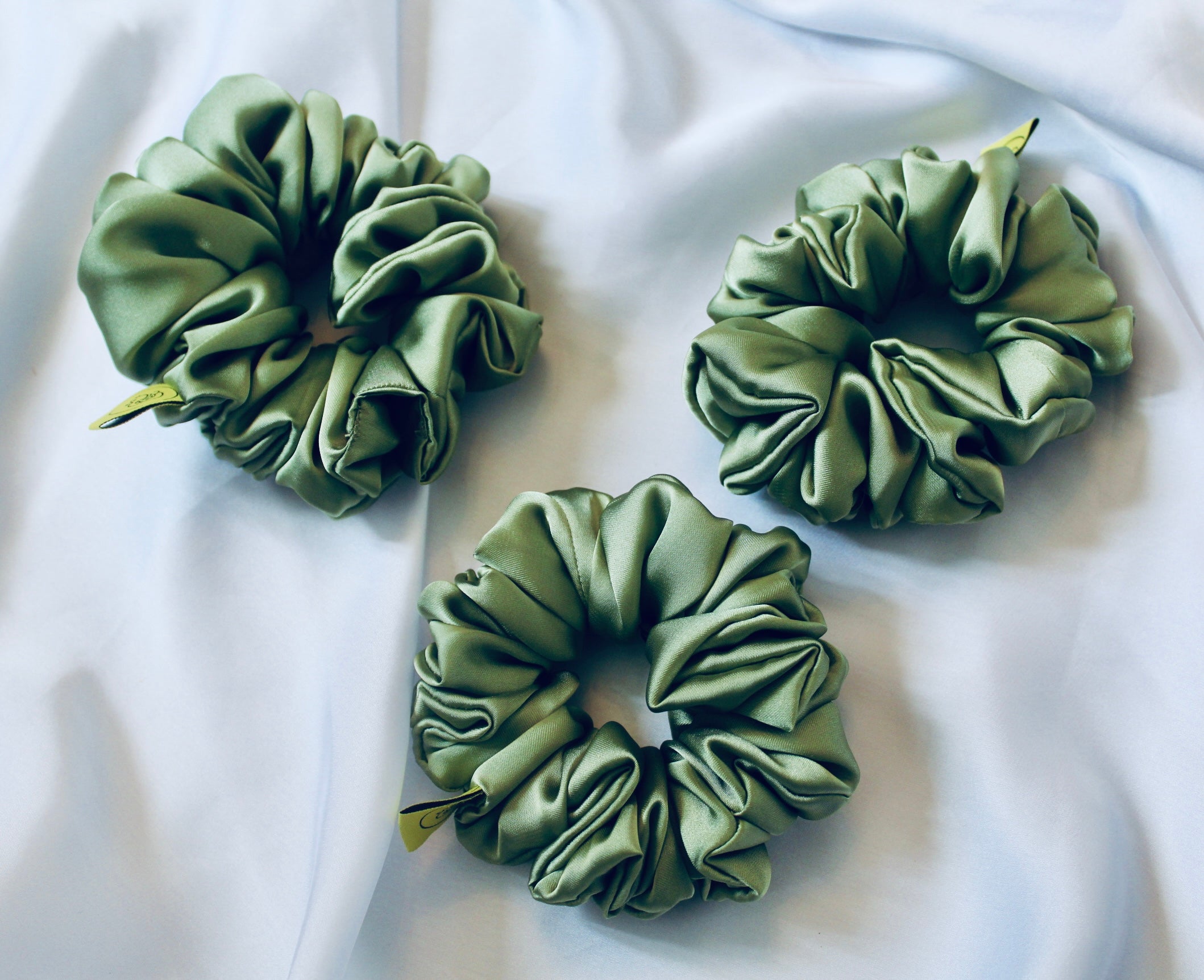 A satin scrunchie in a light green shade with a silky, glossy finish and a ruched design.  Crafted from smooth satin, this scrunchie feels soft and sleek against the hair.  The green tone has a timeless, earthy quality, perfect for both everyday wear and festive styling.  Its ruched fabric is stretched over an elastic band, providing a secure yet comfortable fit.
