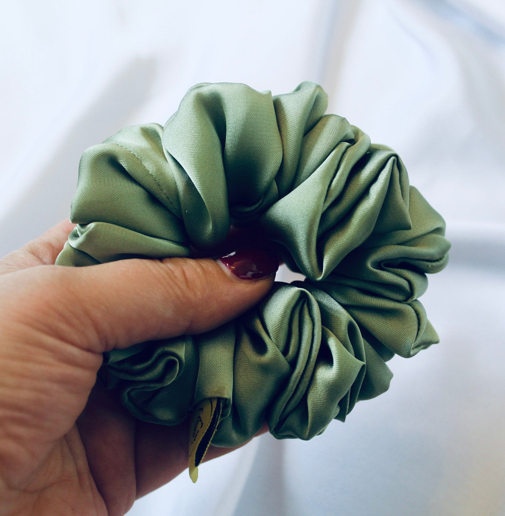 A satin scrunchie in a light green shade with a silky, glossy finish and a ruched design.  Crafted from smooth satin, this scrunchie feels soft and sleek against the hair.  The green tone has a timeless, earthy quality, perfect for both everyday wear and festive styling.  Its ruched fabric is stretched over an elastic band, providing a secure yet comfortable fit.