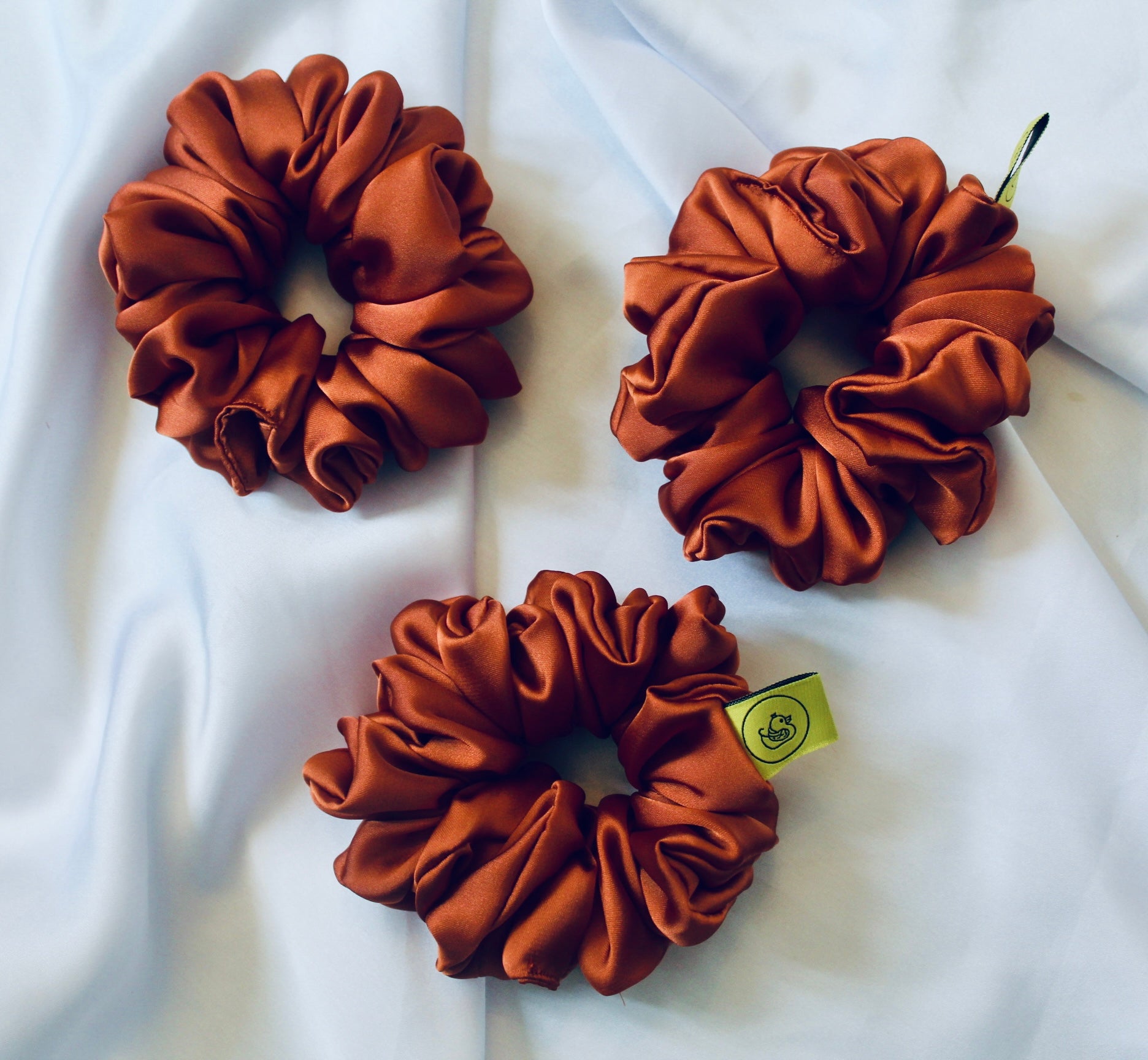 A satin scrunchie in a warm burnt orange shade with a smooth glossy texture and a ruched design.  This scrunchie is made from satin giving it a silky and lightweight feel.  The rich burnt orange colour captures the warmth of autumn leaves, making it perfect seasonal accessory.  The gathered fabric sits on an elastic core, creating a full ruffled look while keeping your hairstyle secure and comfortable.