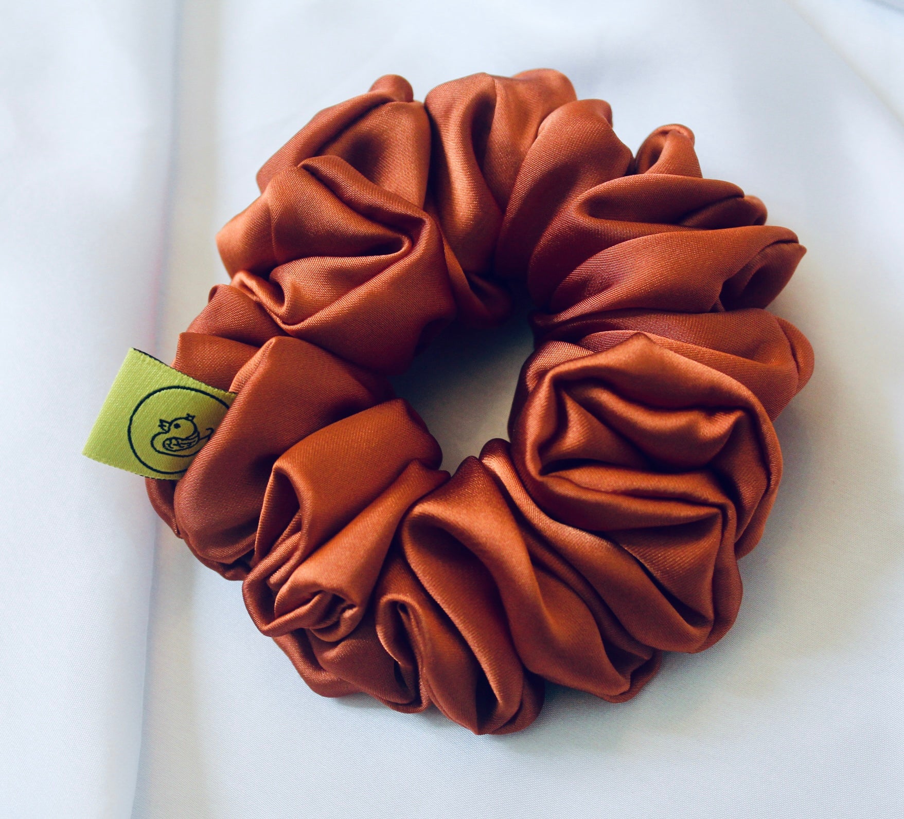 A satin scrunchie in a warm burnt orange shade with a smooth glossy texture and a ruched design.  This scrunchie is made from satin giving it a silky and lightweight feel.  The rich burnt orange colour captures the warmth of autumn leaves, making it perfect seasonal accessory.  The gathered fabric sits on an elastic core, creating a full ruffled look while keeping your hairstyle secure and comfortable.