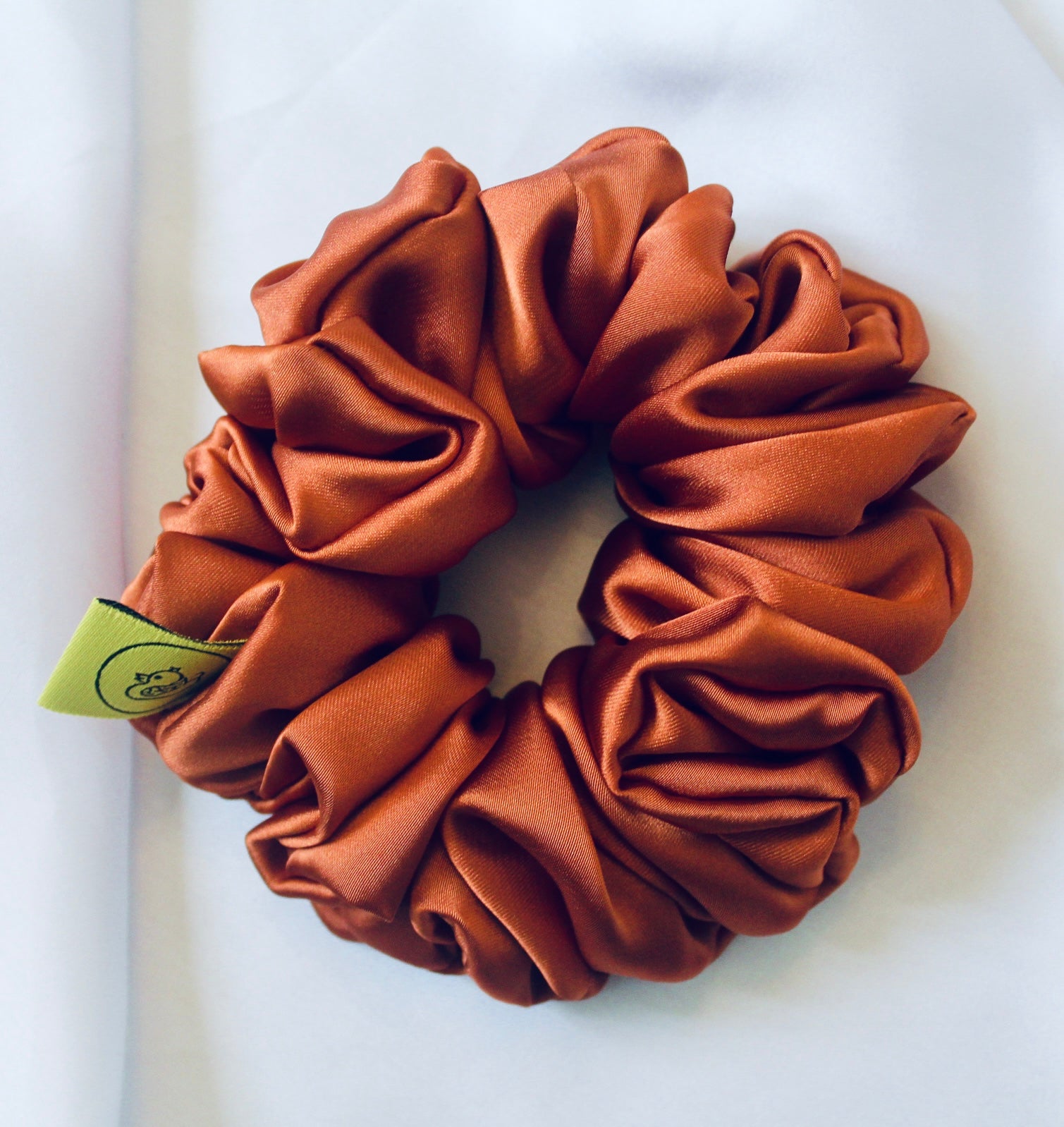 A satin scrunchie in a warm burnt orange shade with a smooth glossy texture and a ruched design.  This scrunchie is made from satin giving it a silky and lightweight feel.  The rich burnt orange colour captures the warmth of autumn leaves, making it perfect seasonal accessory.  The gathered fabric sits on an elastic core, creating a full ruffled look while keeping your hairstyle secure and comfortable.